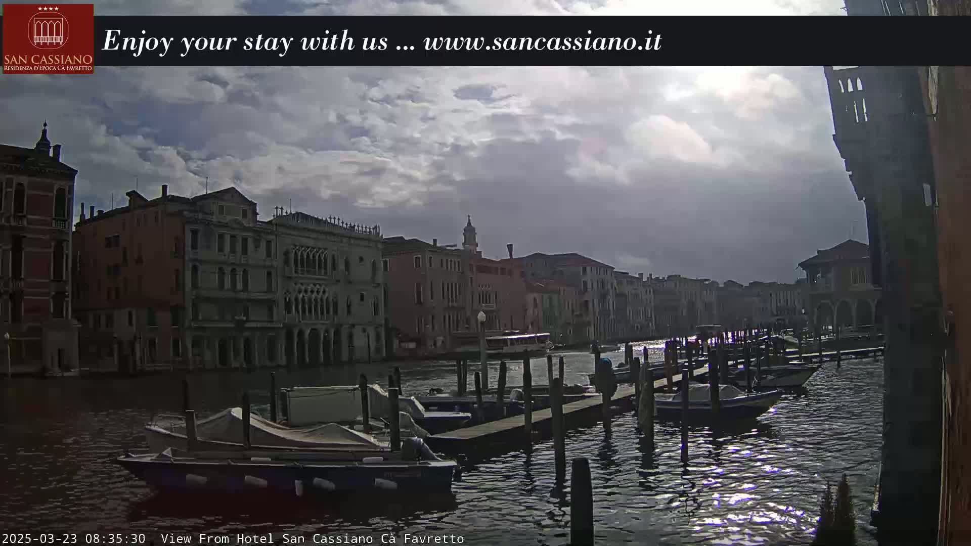 A cloudy day in Venice shows several moored boats along a canal lined with buildings.