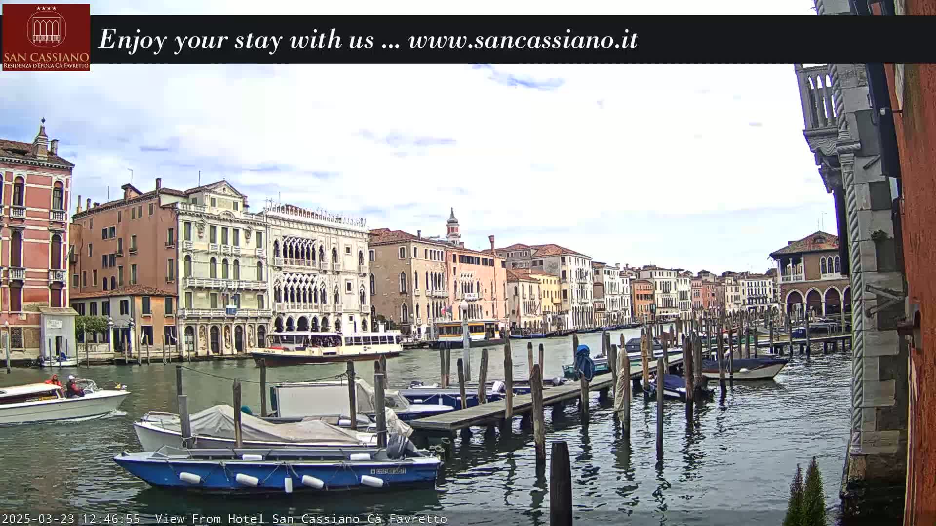 A canal in Venice, Italy, with several boats moored along a wooden dock, is viewed under partly cloudy skies.