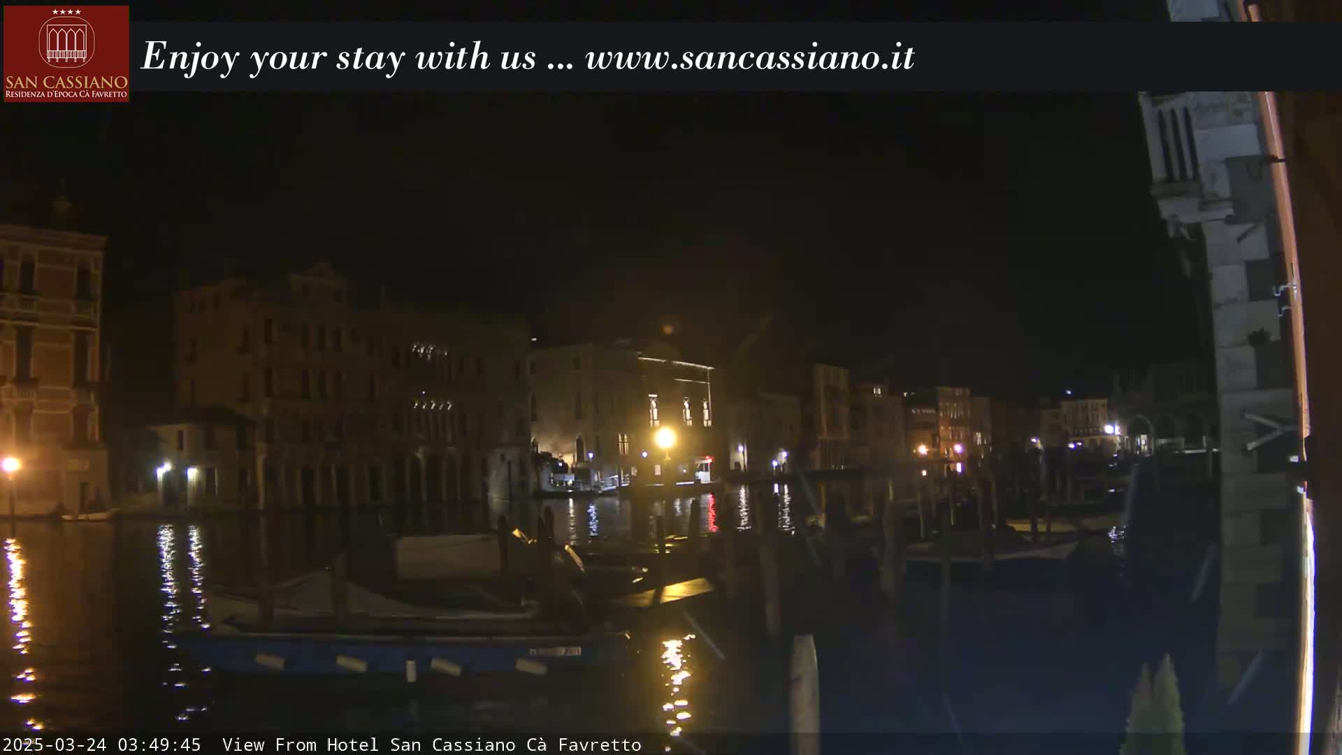 A nighttime view across a canal shows several buildings lit along the water's edge, with boats moored nearby.