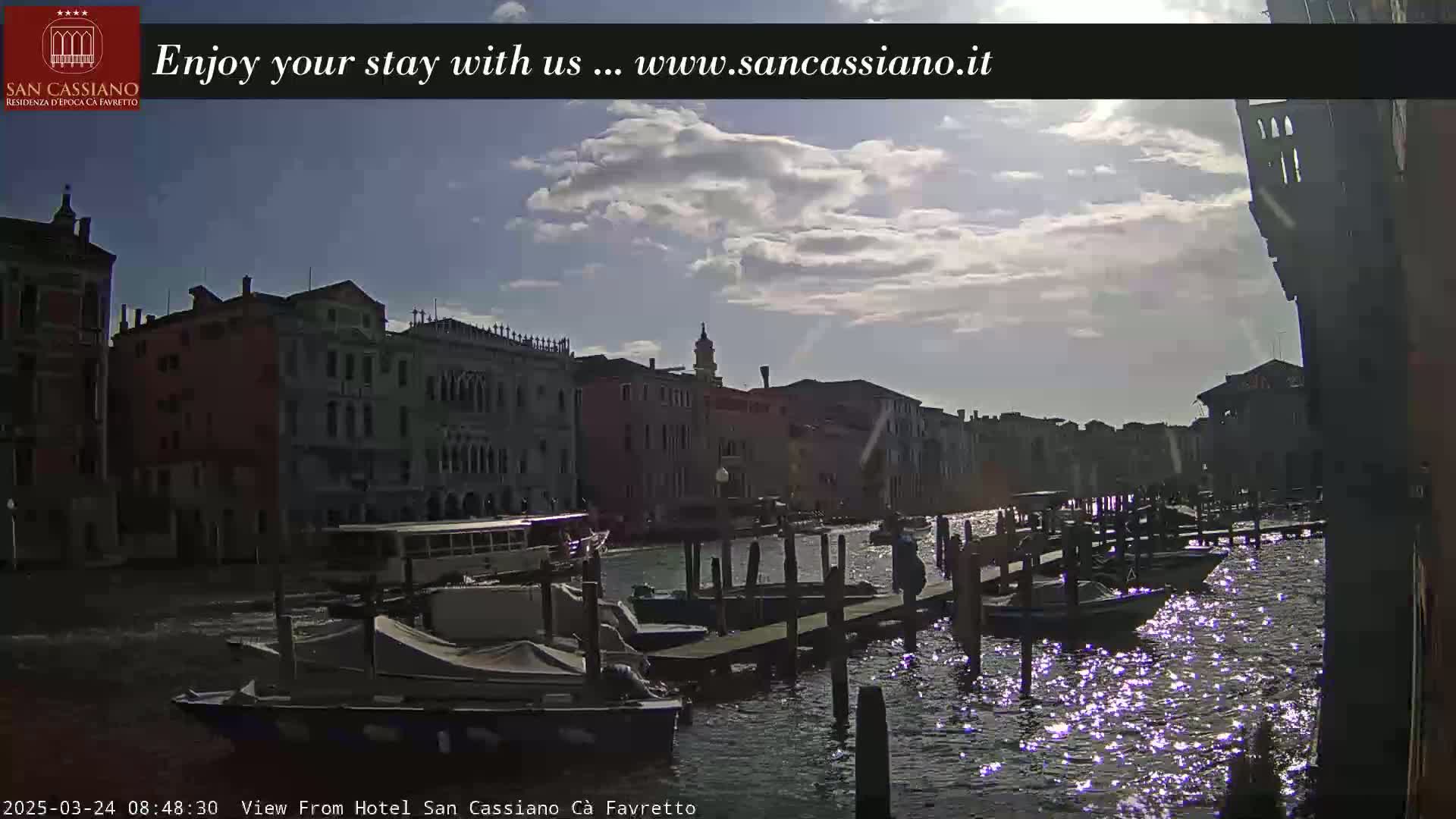 A canal in Venice, under a partly cloudy sky, shows several boats moored at wooden docks beside buildings along the waterway.