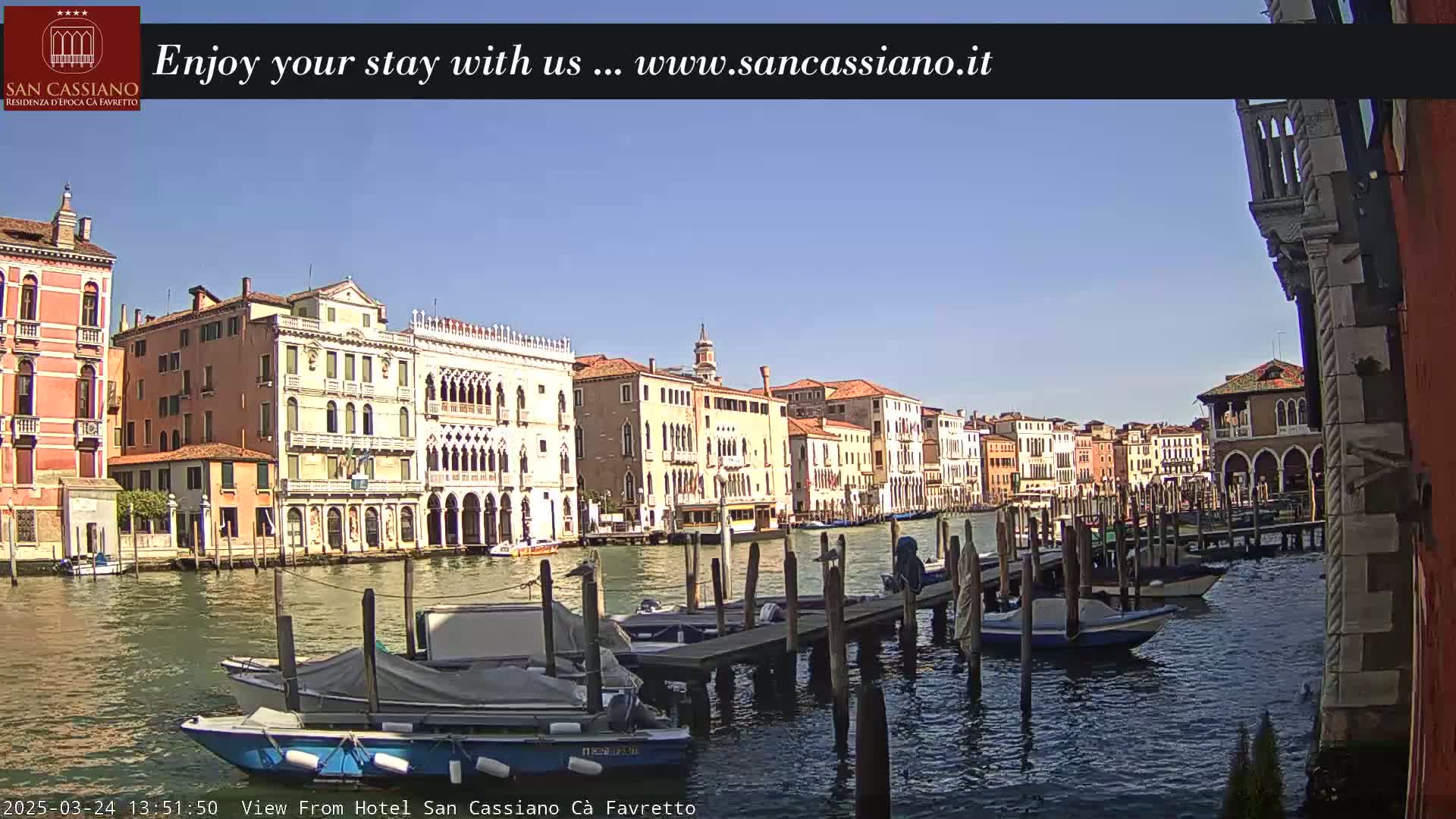 A sunny day in Venice shows a canal lined with colorful buildings, boats moored at wooden docks, and calm water reflecting the architecture.