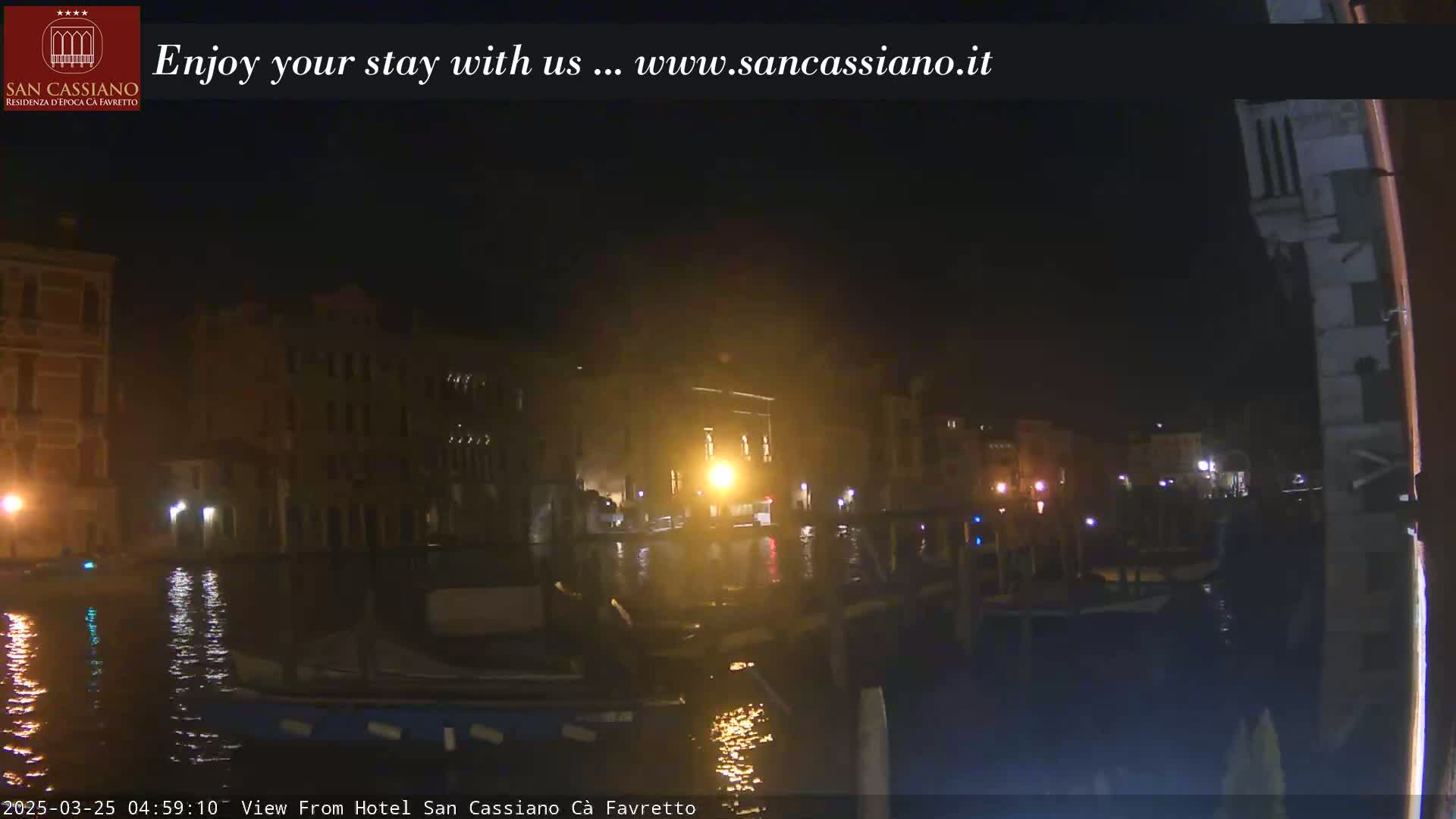 A nighttime view of a canal in Venice, showing dark buildings with some lights visible along the water.