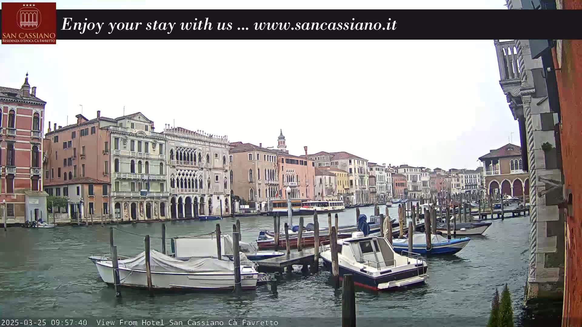 A canal in Venice, Italy, with numerous boats moored along the sides, under an overcast sky.