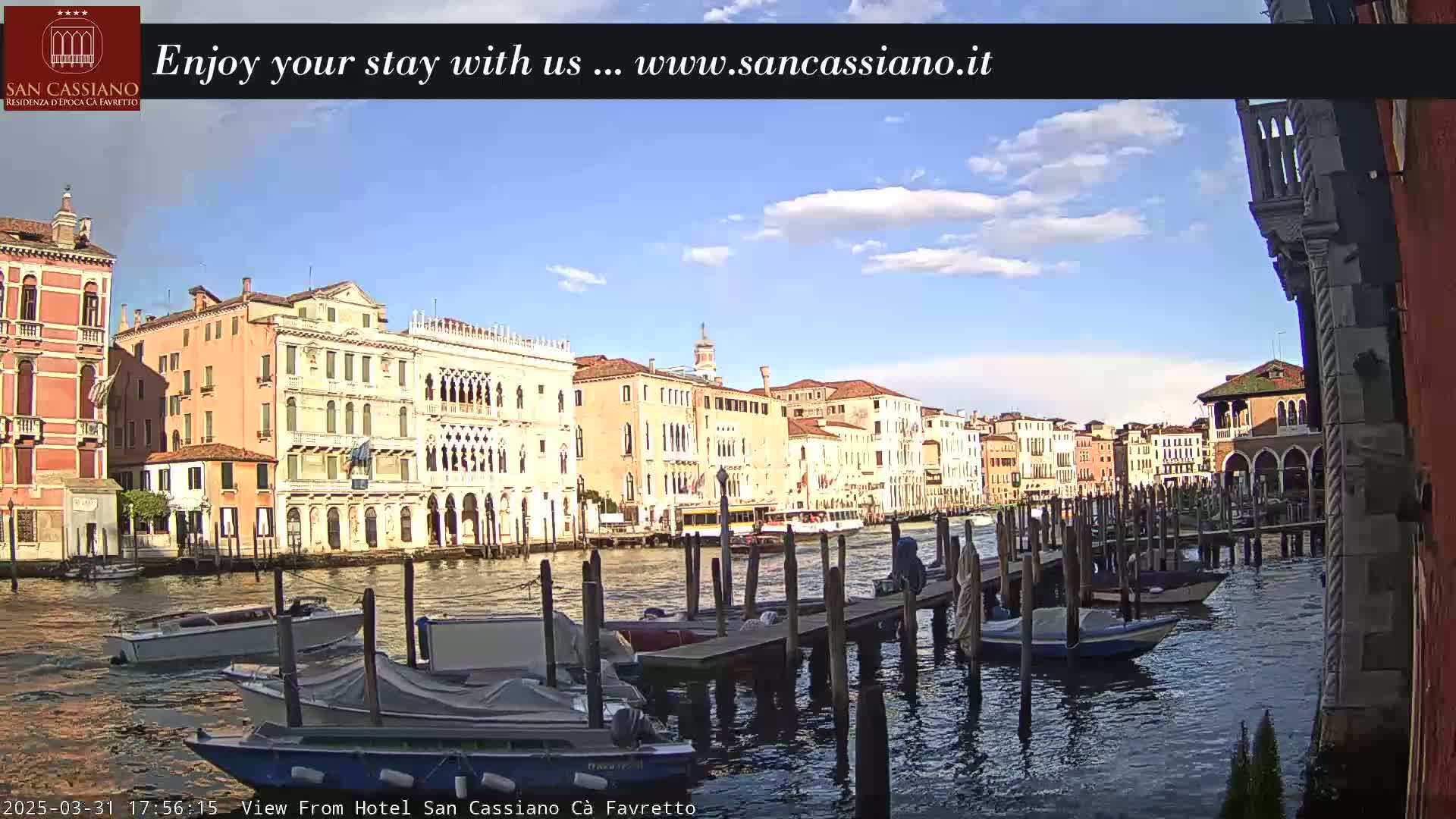 A sunny day in Venice shows colorful buildings lining a canal, with several boats moored at a dock.