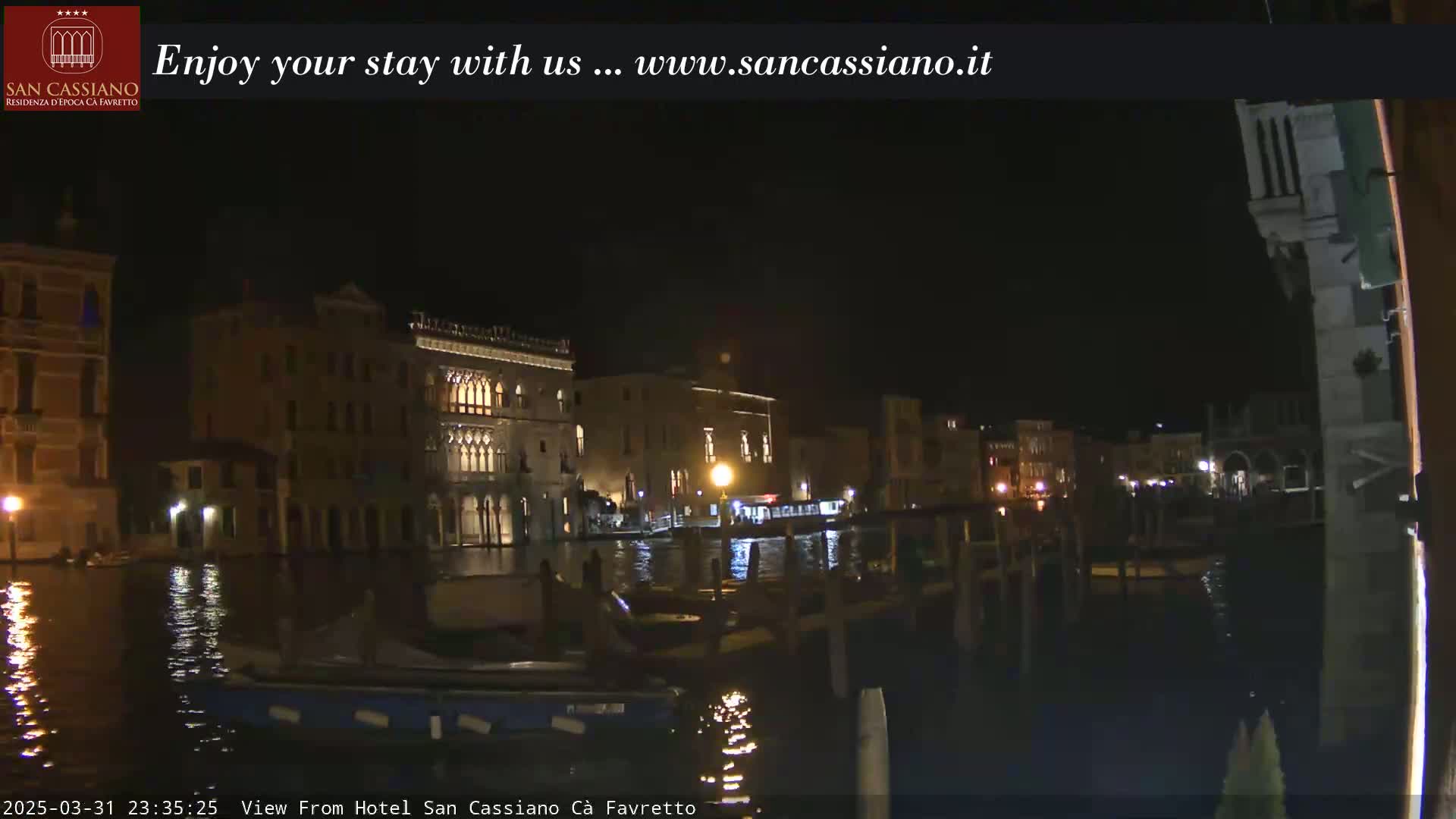 A nighttime view of illuminated buildings along a canal, possibly in Venice, with calm water reflecting the lights.