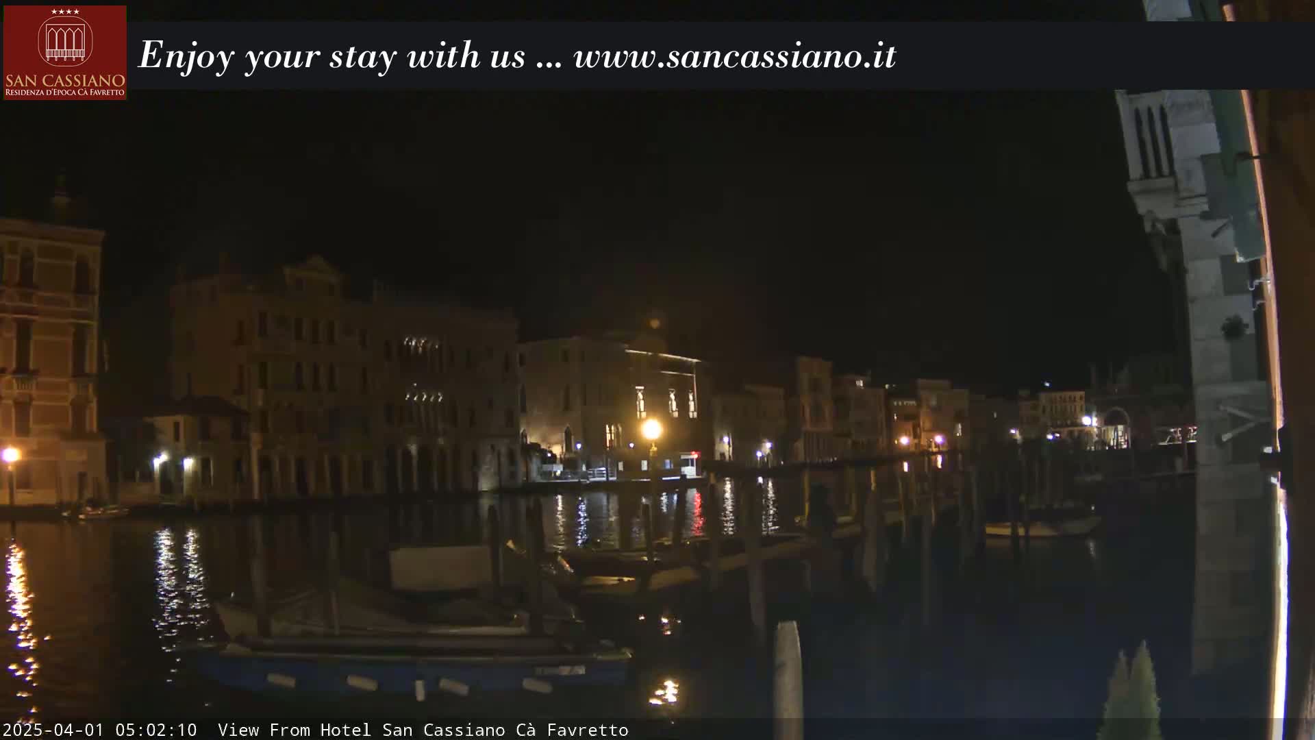 A nighttime view of a canal in Venice, with illuminated buildings lining the waterway, reflecting softly in the dark water.