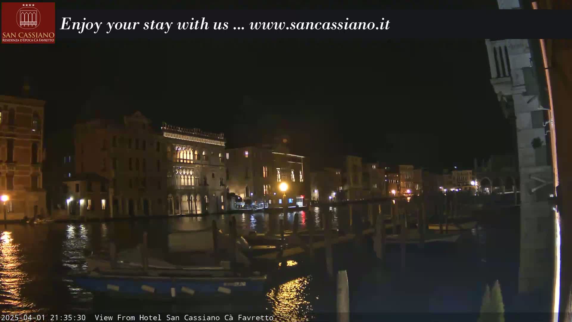 A nighttime view of illuminated buildings along a canal, with boats moored at the waterfront under a clear, dark sky.