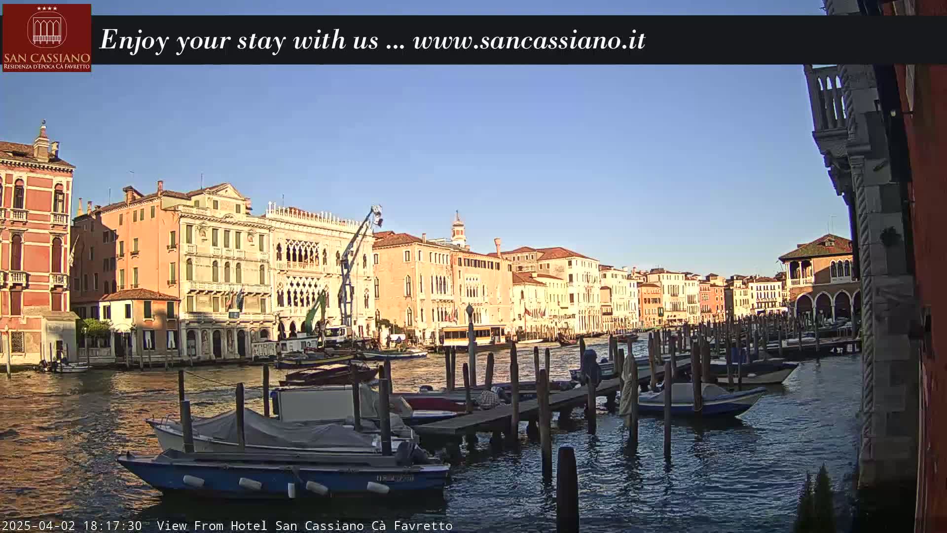 A canal in Venice, Italy, with numerous boats moored along a dock, under a clear blue sky at sunset.