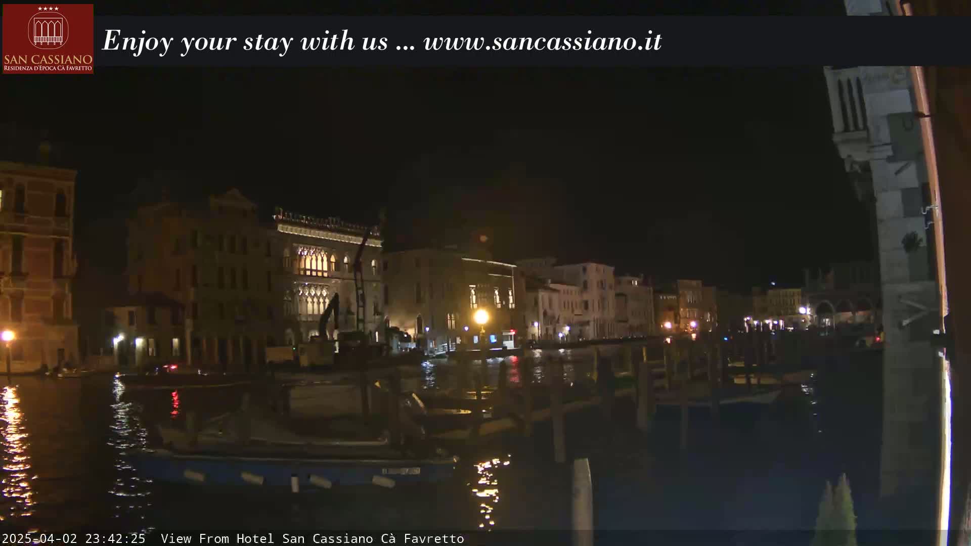 A nighttime view of a canal in Venice, showing illuminated buildings and boats on the water under a dark sky.