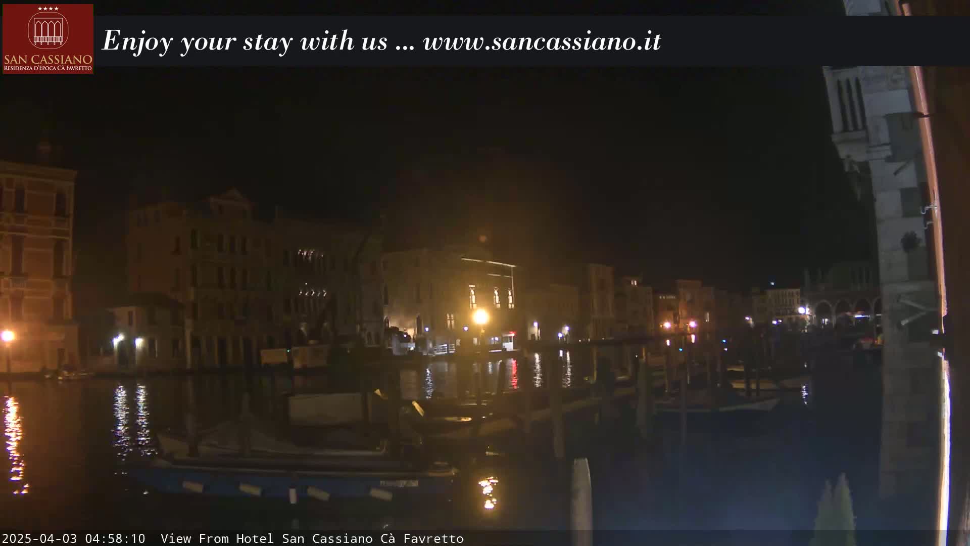 A nighttime view of a canal in Venice, Italy, shows buildings lit along the waterway, with boats moored along the edge.