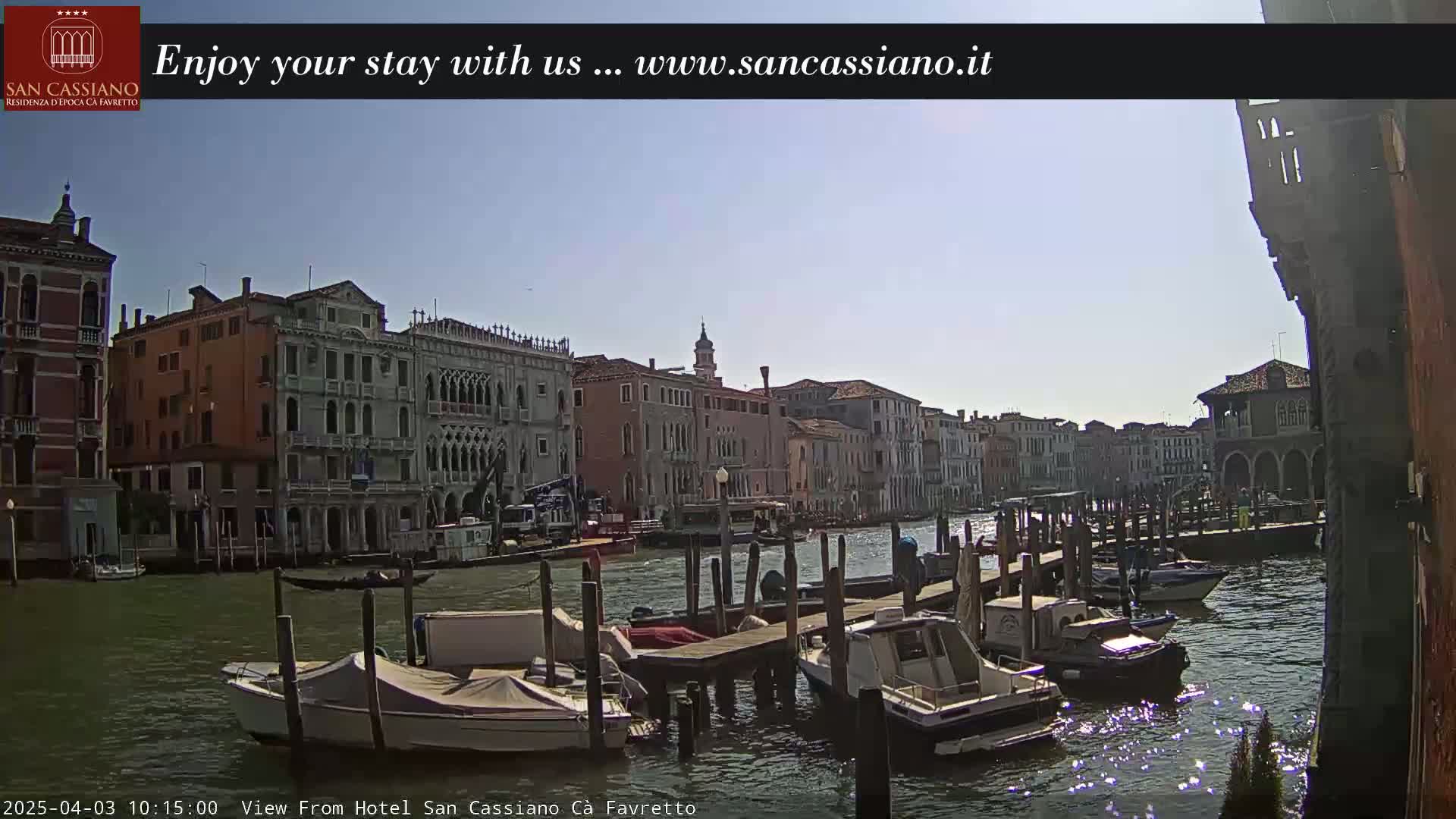 A sunny day in Venice shows a canal with several moored boats and numerous historic buildings lining the waterway.