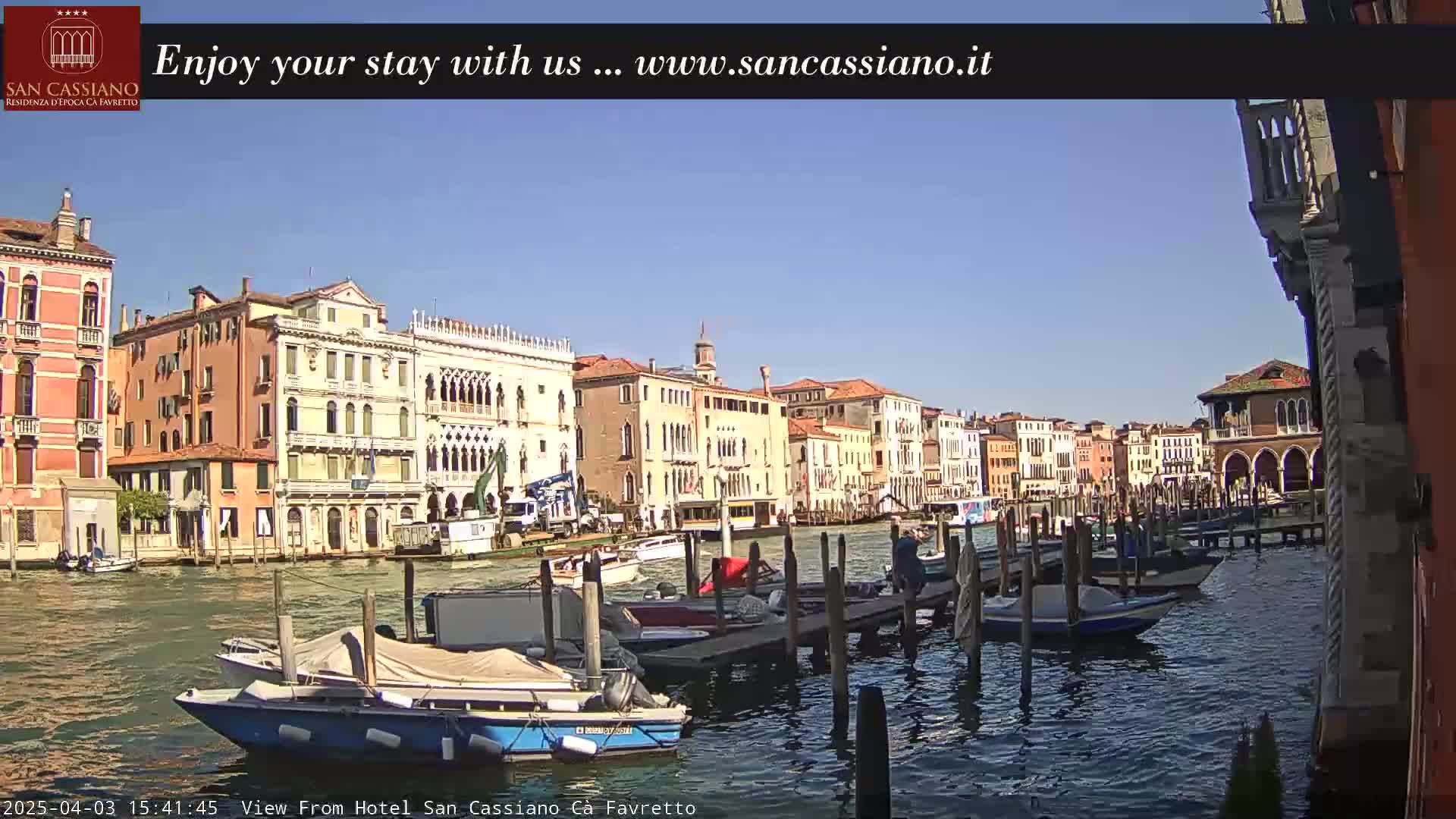 A sunny day in Venice shows colorful buildings lining a canal, with several boats moored at a dock.