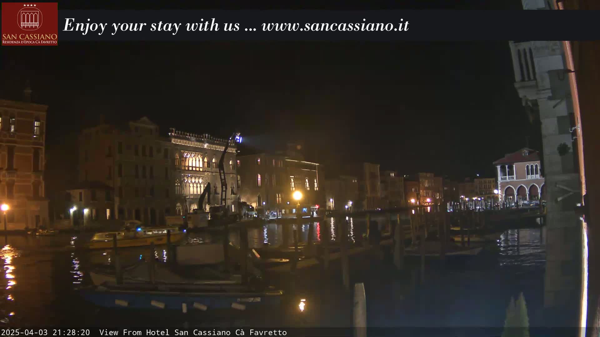 A nighttime view of a canal in Venice, with illuminated buildings lining the waterway and boats moored along the edges.