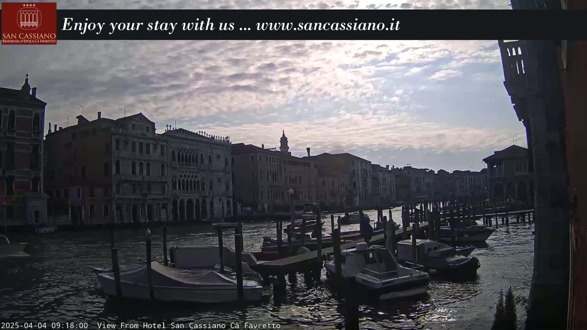 Numerous boats are moored along a canal beside historic buildings under a partly cloudy sky.