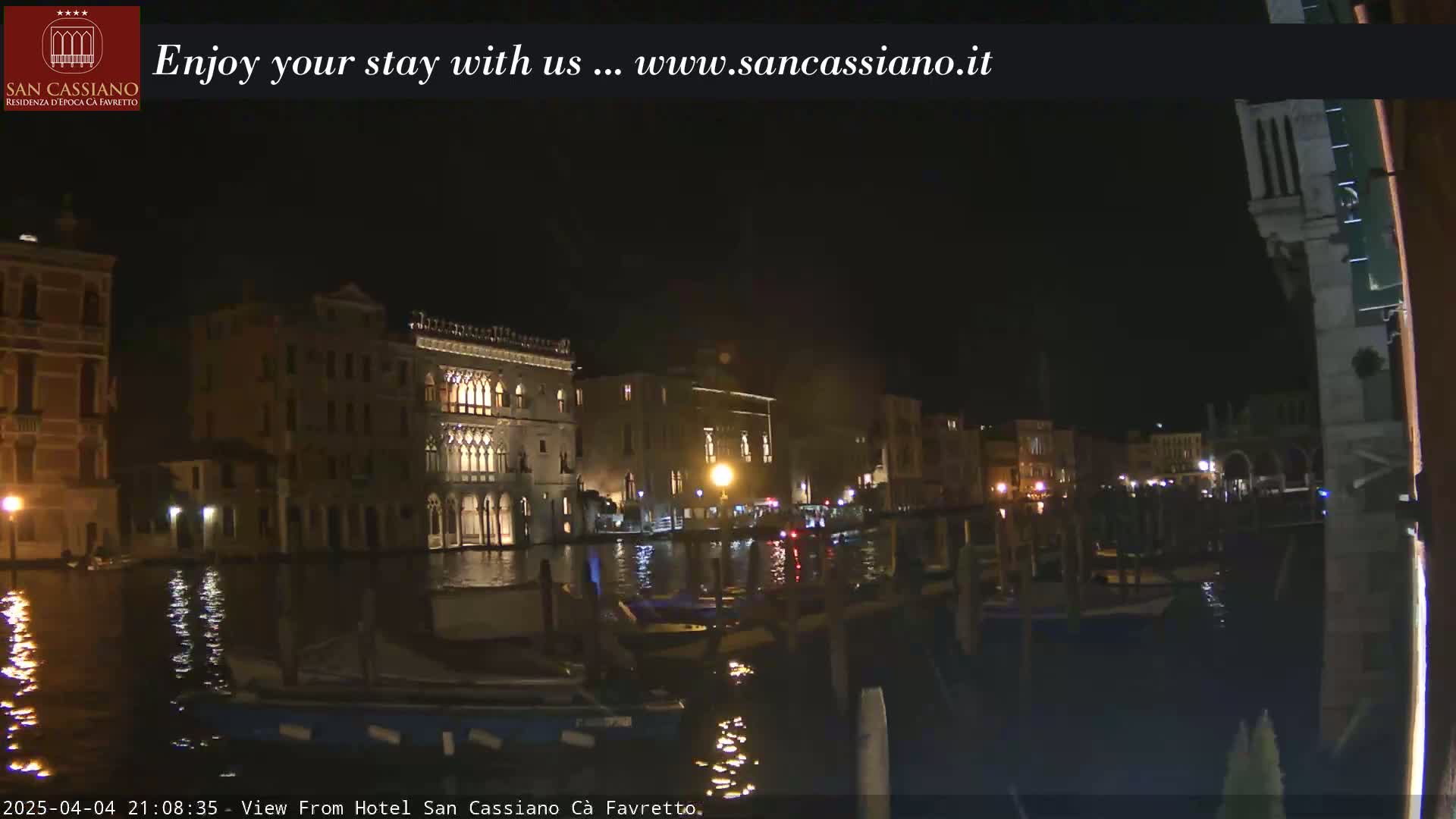 A nighttime view of a canal in Venice, Italy, with illuminated buildings lining the waterway and several boats moored along the edges.