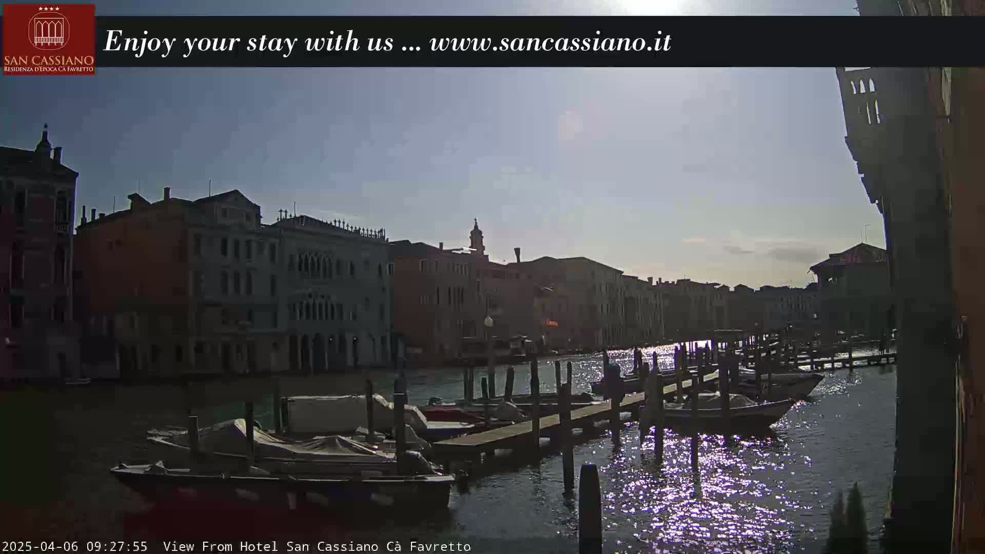 A sunny day in Venice shows a canal lined with docked boats and buildings, with the sun reflecting on the water.