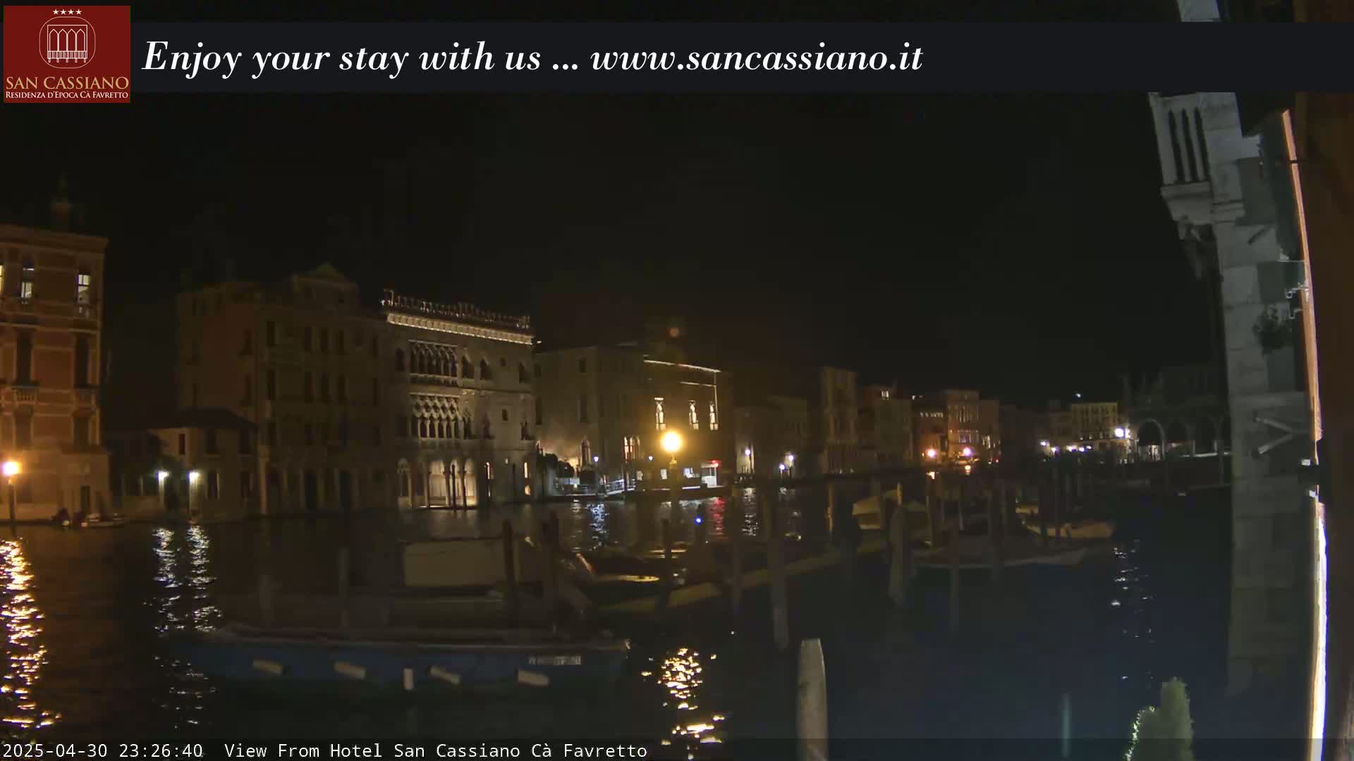 A nighttime view of a canal in Venice, showing illuminated buildings along the waterway and several moored boats.