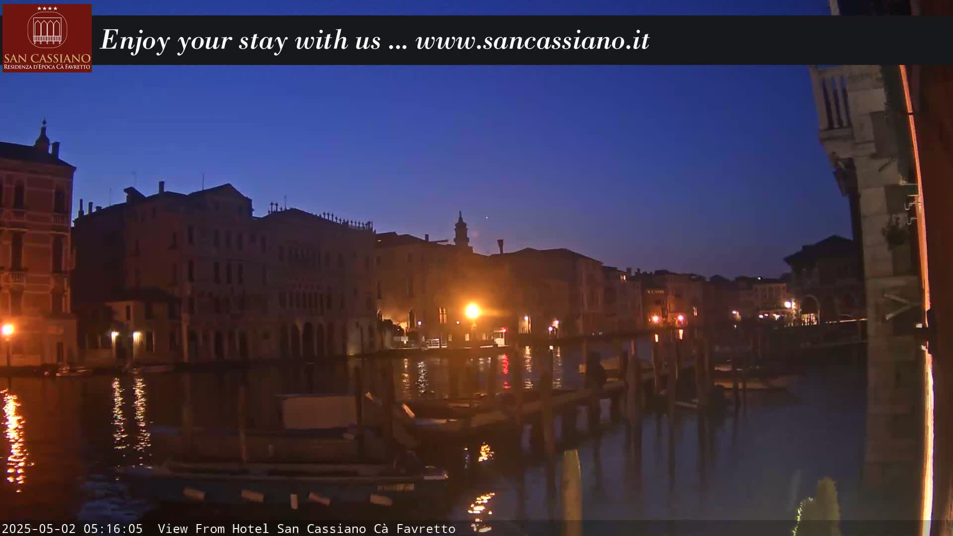 A twilight view of a canal in Venice, showing tall buildings lit by a few warm lights against a dark blue sky.