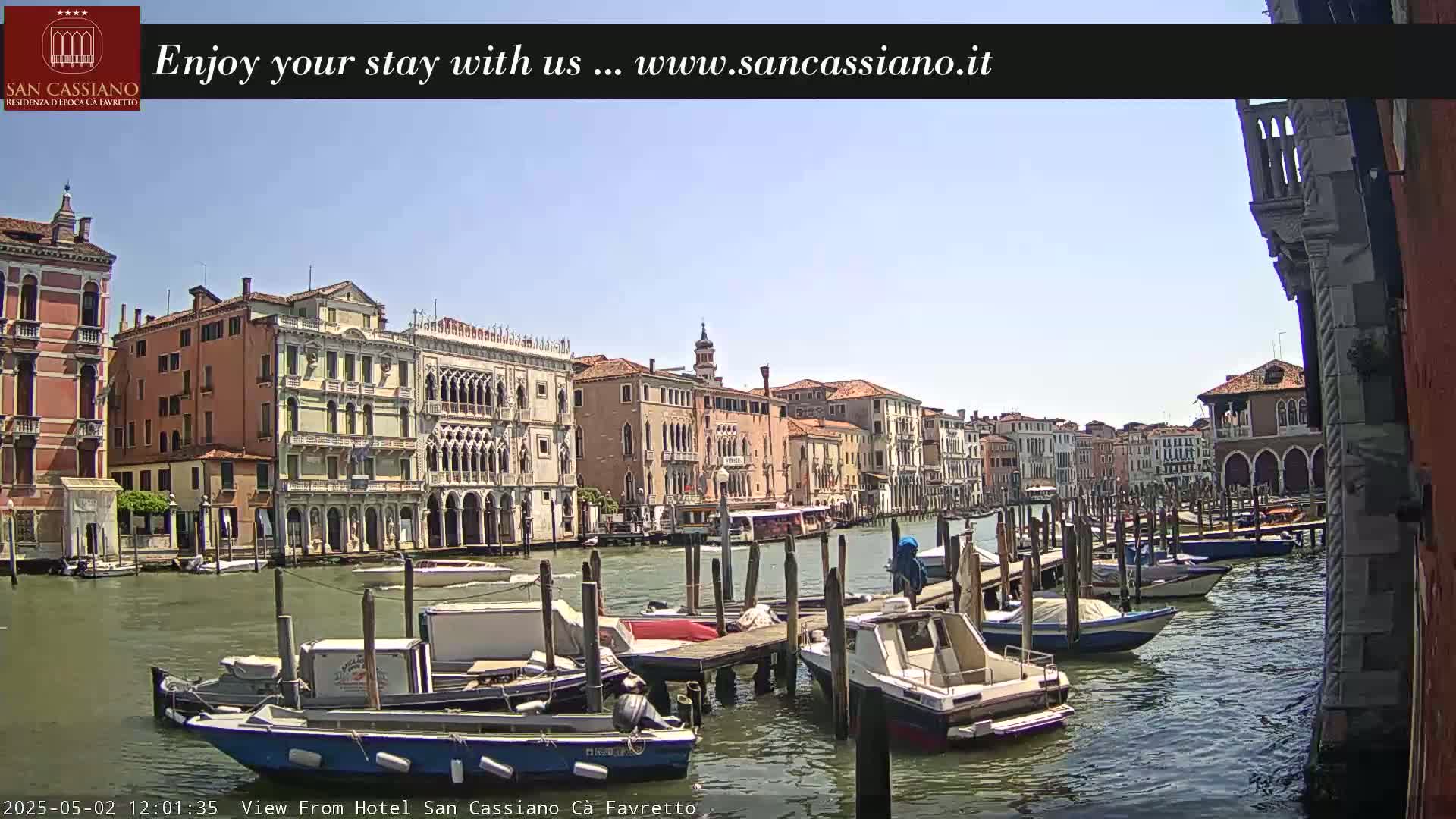 A sunny day in Venice shows a canal lined with buildings, boats docked at piers, and a water bus.