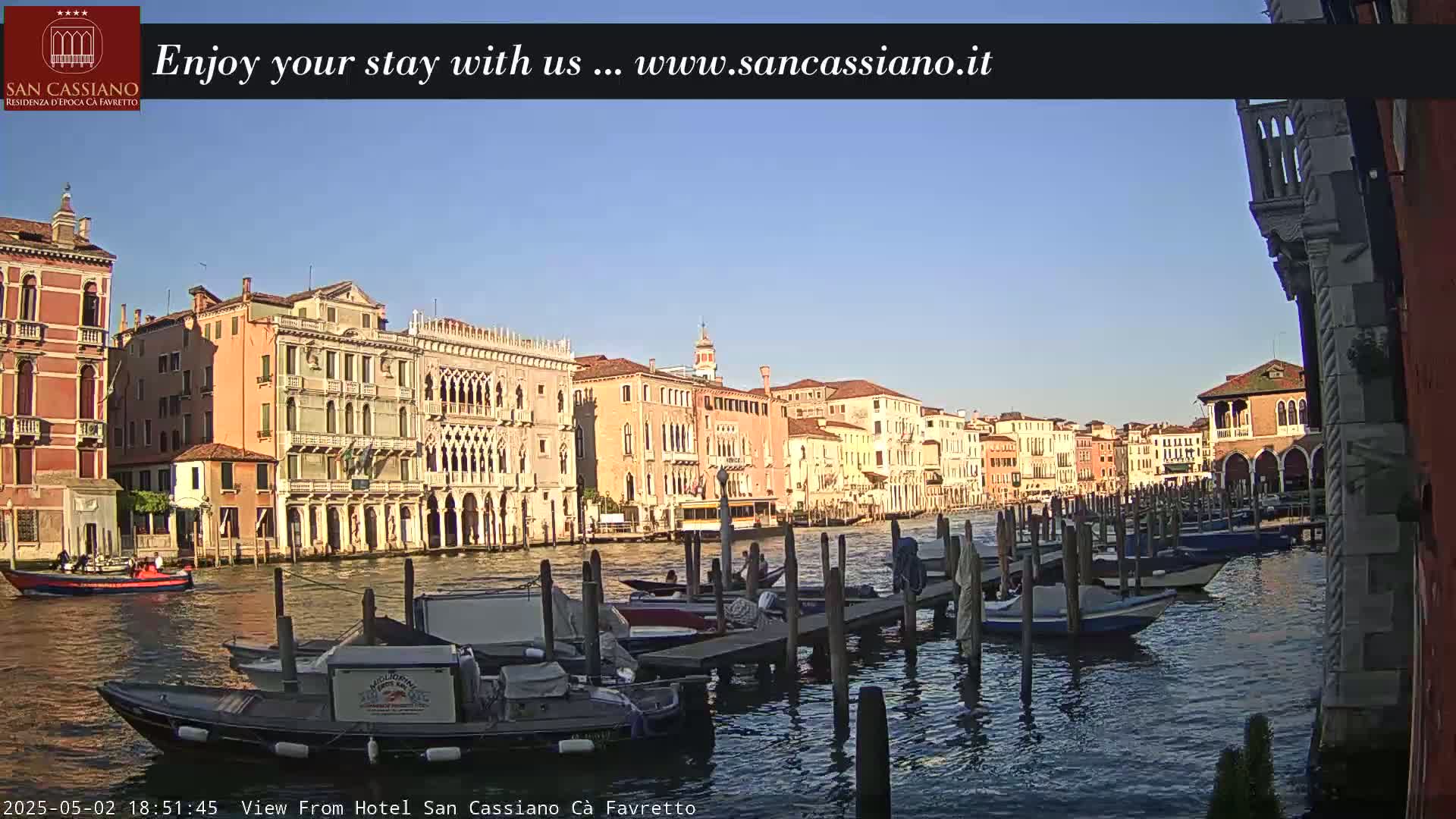 Under a clear blue sky, a canal in Venice is lined with colorful buildings and numerous boats moored at wooden docks.