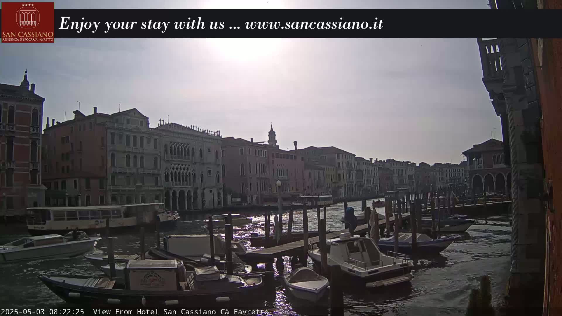 A sunny day in Venice shows a canal with numerous boats moored along the waterfront, in front of several tall buildings.
