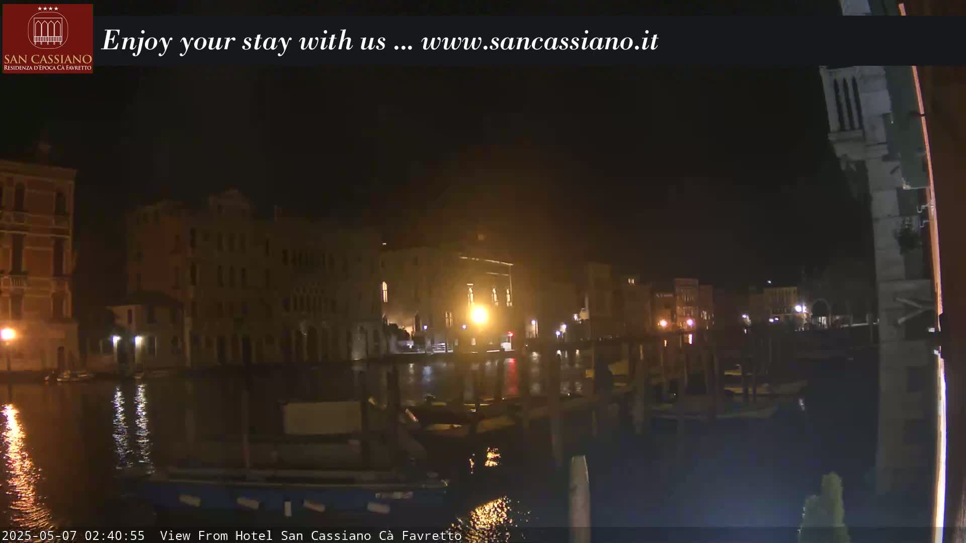 A nighttime view across a canal shows illuminated buildings on both sides, with boats moored along the waterfront.