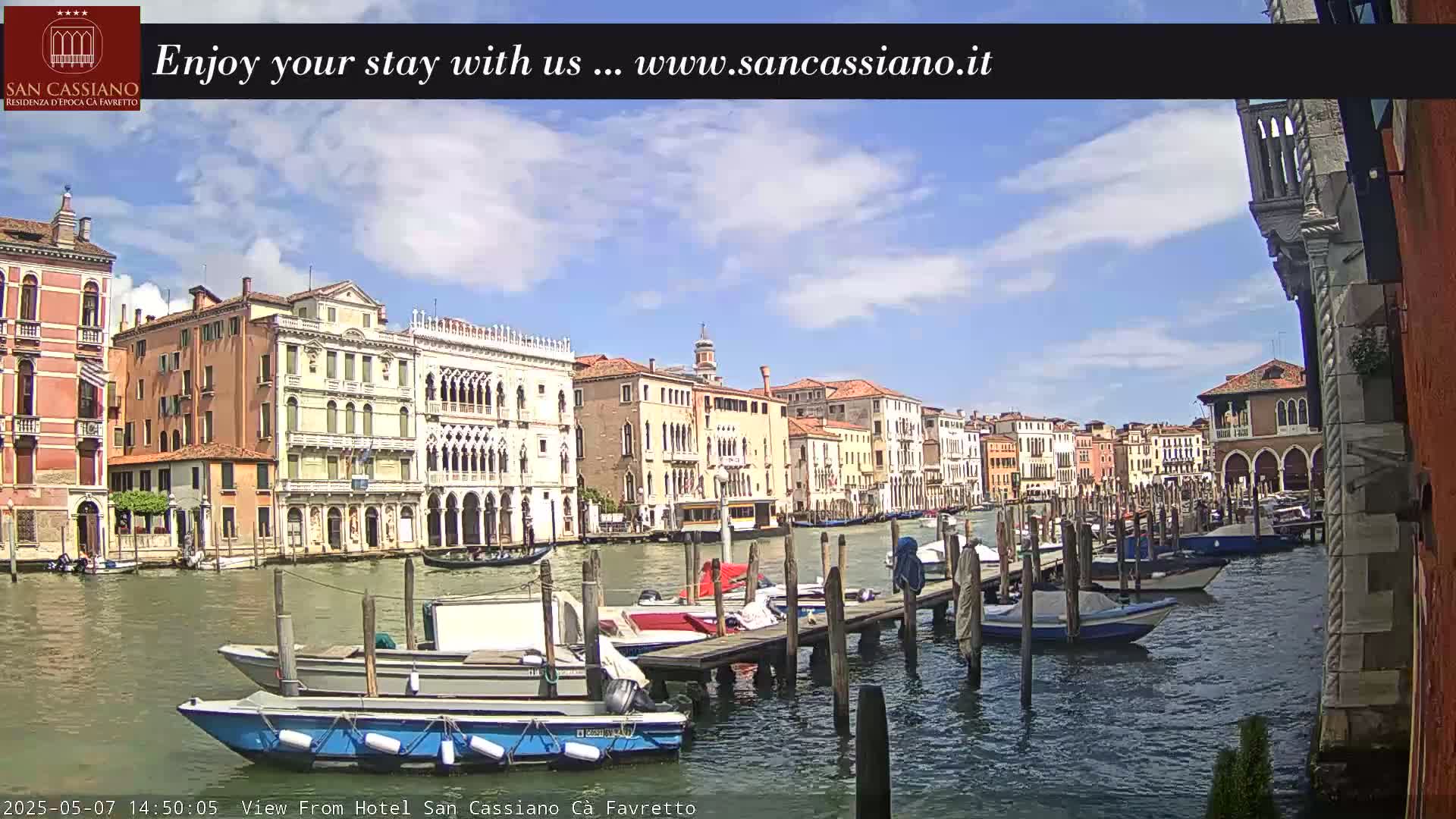 A sunny day in Venice shows a canal lined with colorful buildings and numerous boats moored at wooden docks.