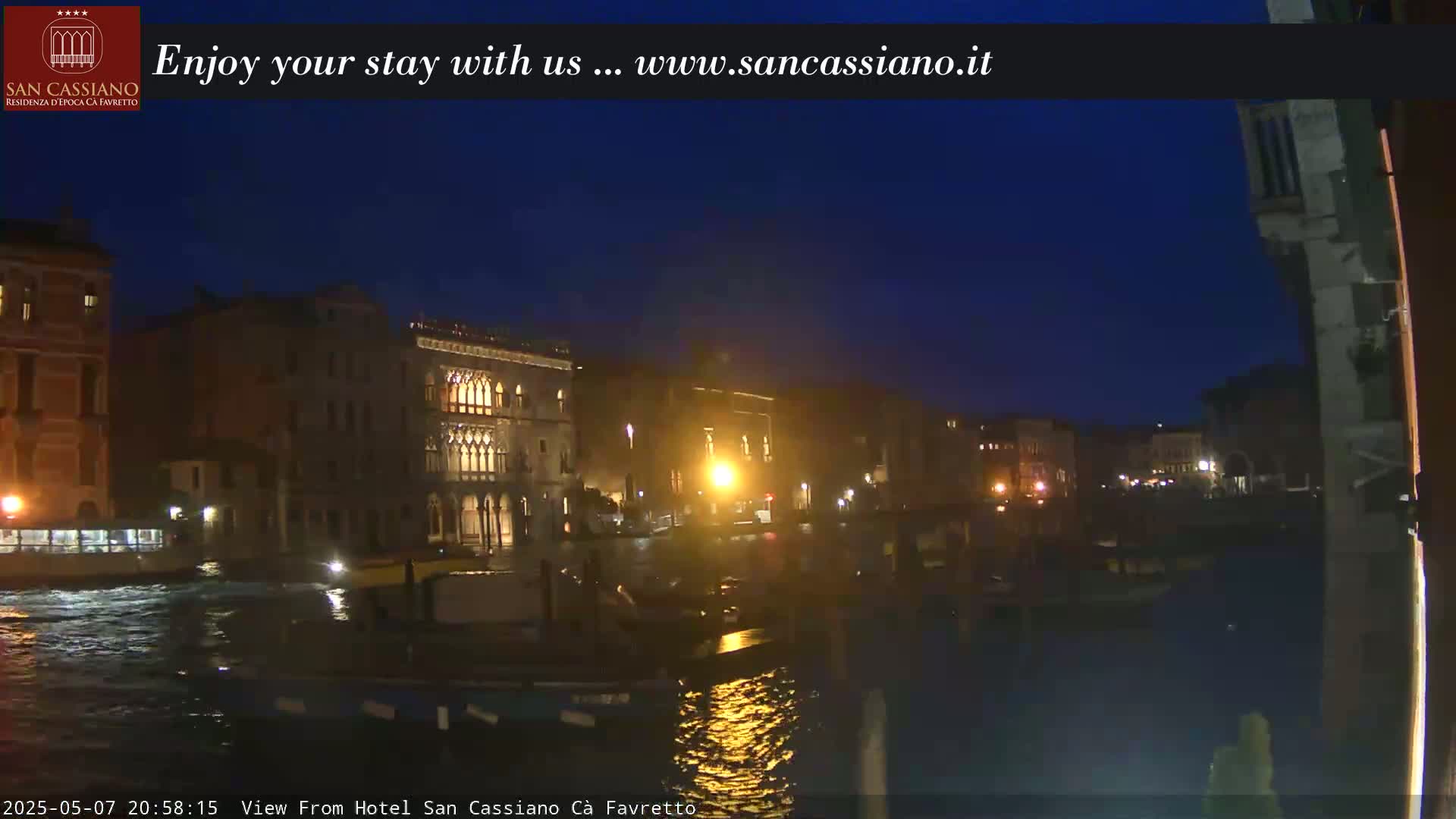 A nighttime view of illuminated buildings along a waterway, under a dark blue sky.