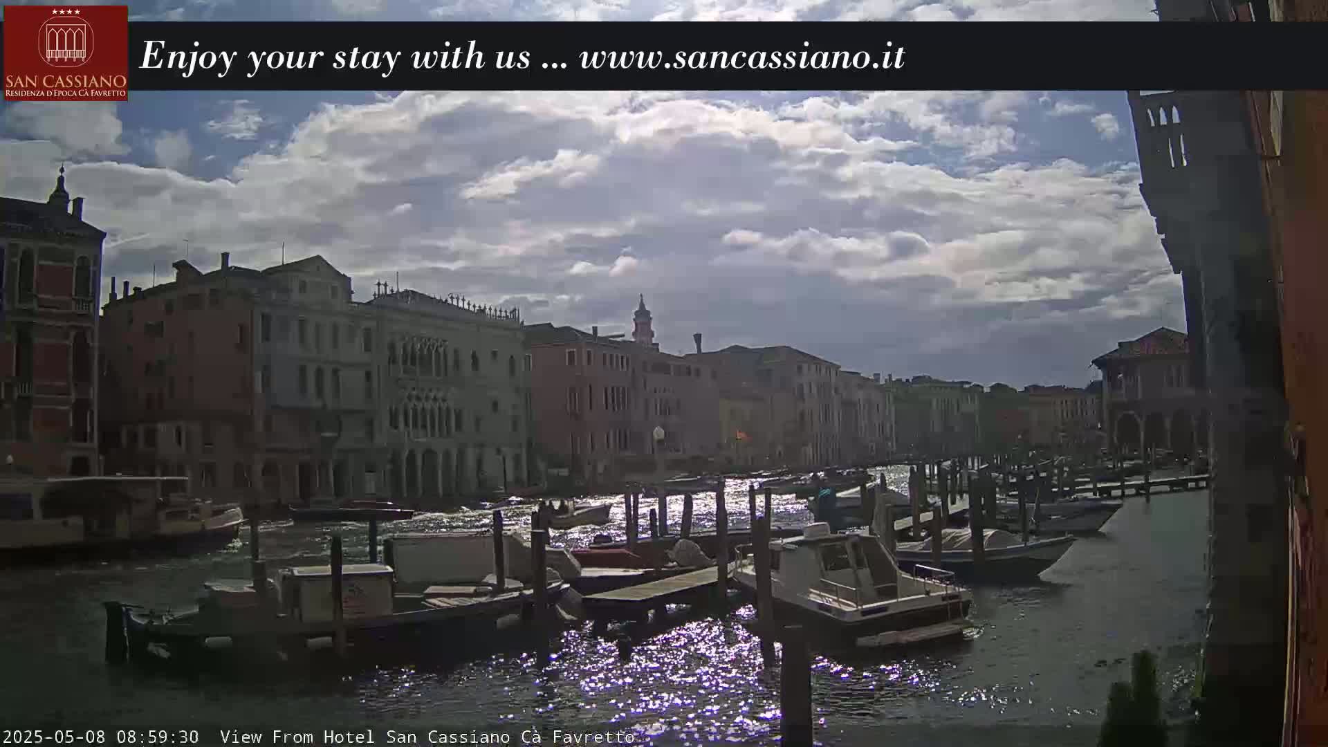 A partly cloudy sky overlooks a Venetian canal, where several boats are moored along the waterway, with buildings lining both sides.