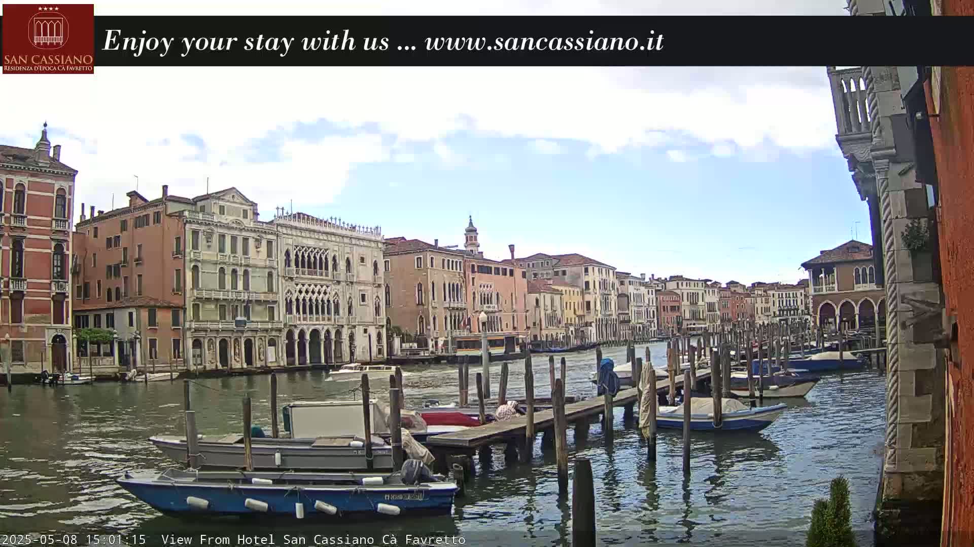 A partly cloudy day shows a Venetian canal with several boats moored at wooden docks, beside buildings along the waterway.