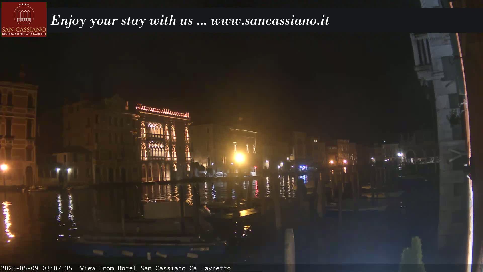 A nighttime view across a calm canal shows illuminated buildings along the waterfront.