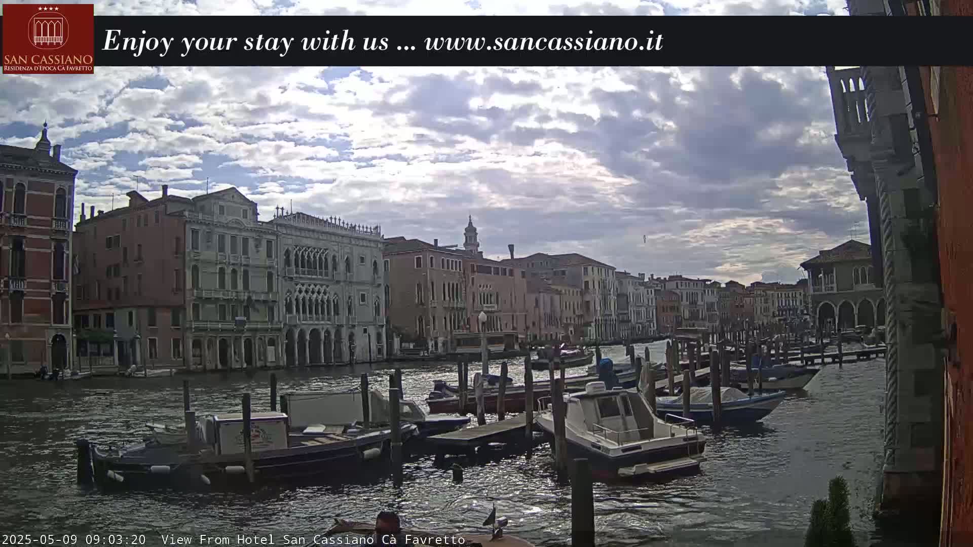 A canal in Venice, under a partly cloudy sky, is lined with historic buildings and numerous moored boats.