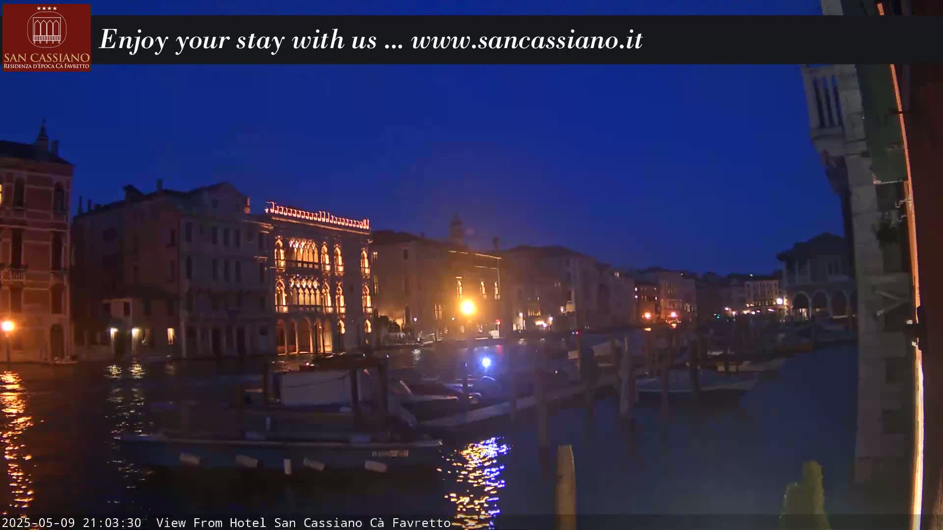 Illuminated Venetian buildings line a canal at twilight, with several boats moored along the waterway.