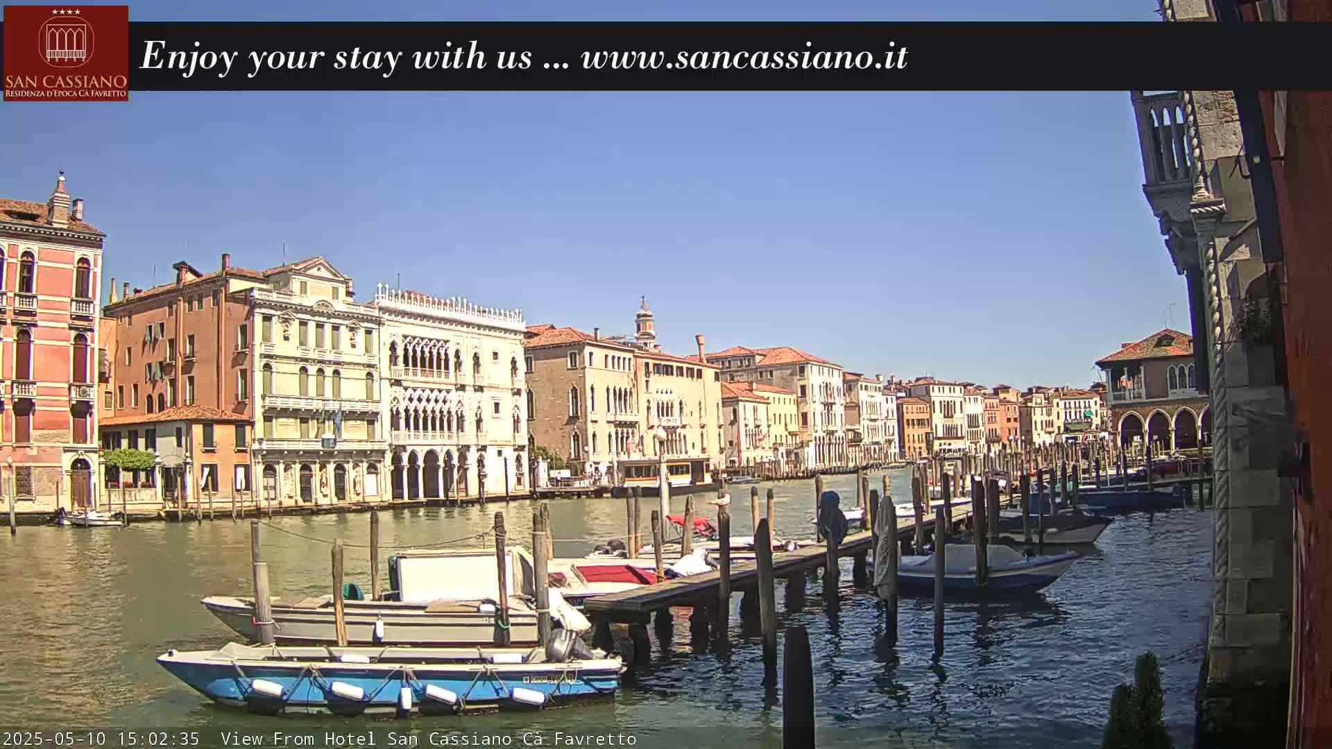 Under a clear blue sky, several boats are moored at a dock along a canal lined with Venetian buildings.