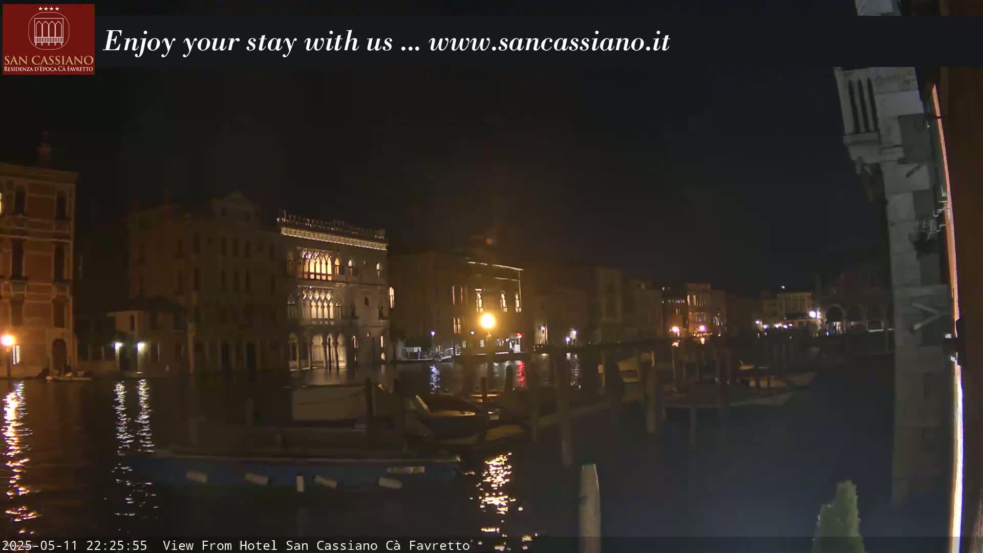 Nighttime view of illuminated buildings along a canal, with several boats moored nearby.