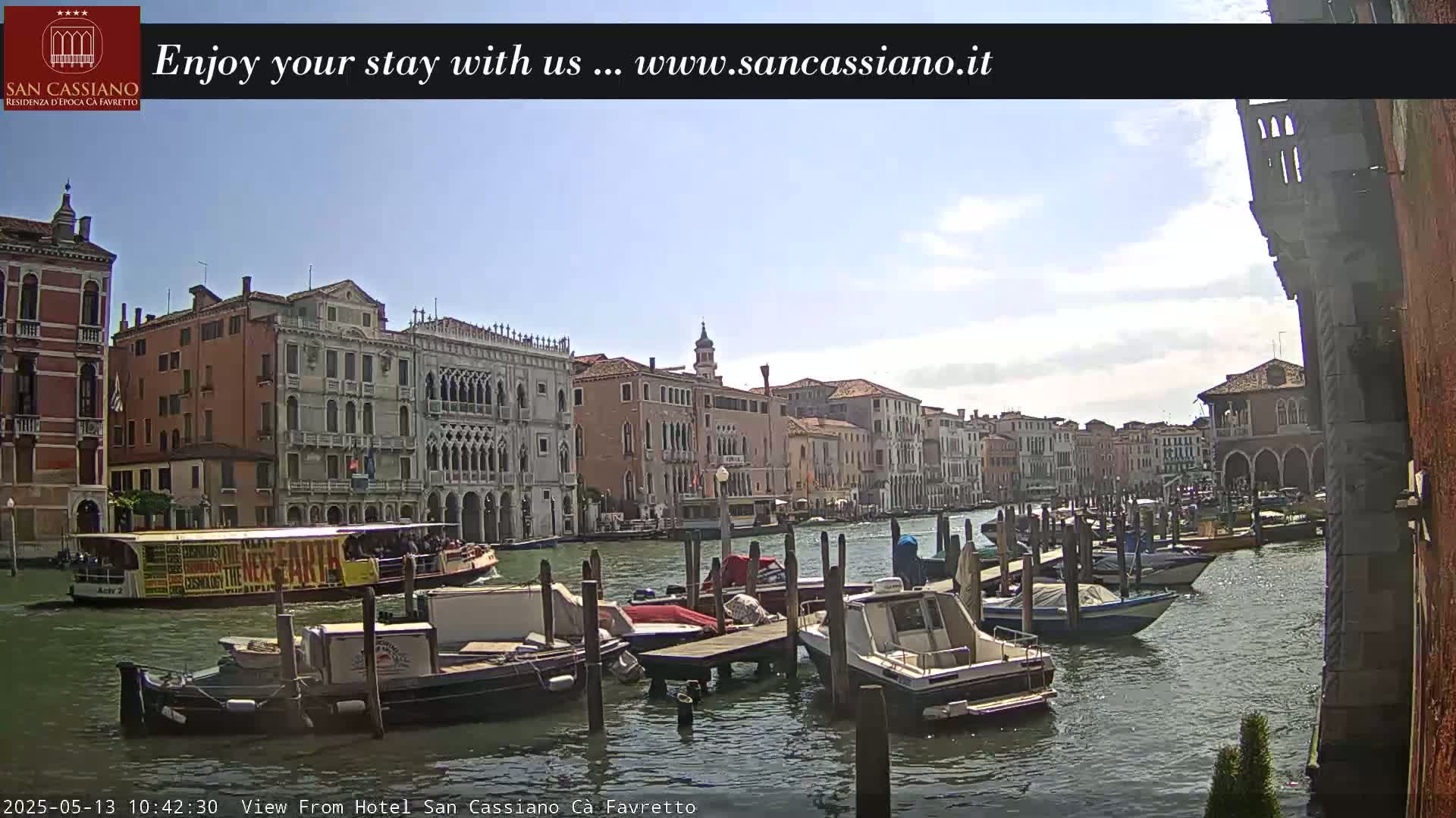 A sunny day in Venice shows a canal lined with historic buildings and numerous boats moored at docks.