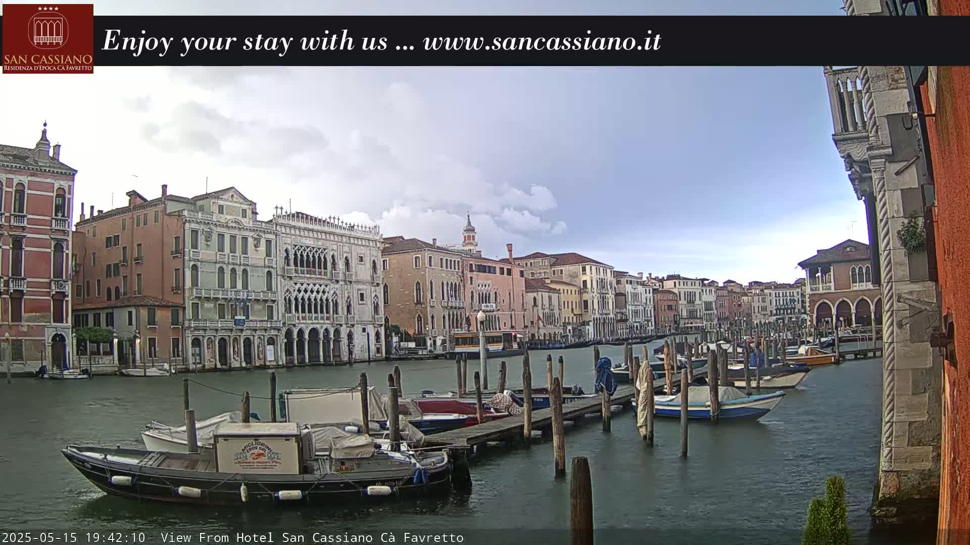 A canal in Venice, under a partly cloudy sky, is lined with historic buildings and numerous moored boats.