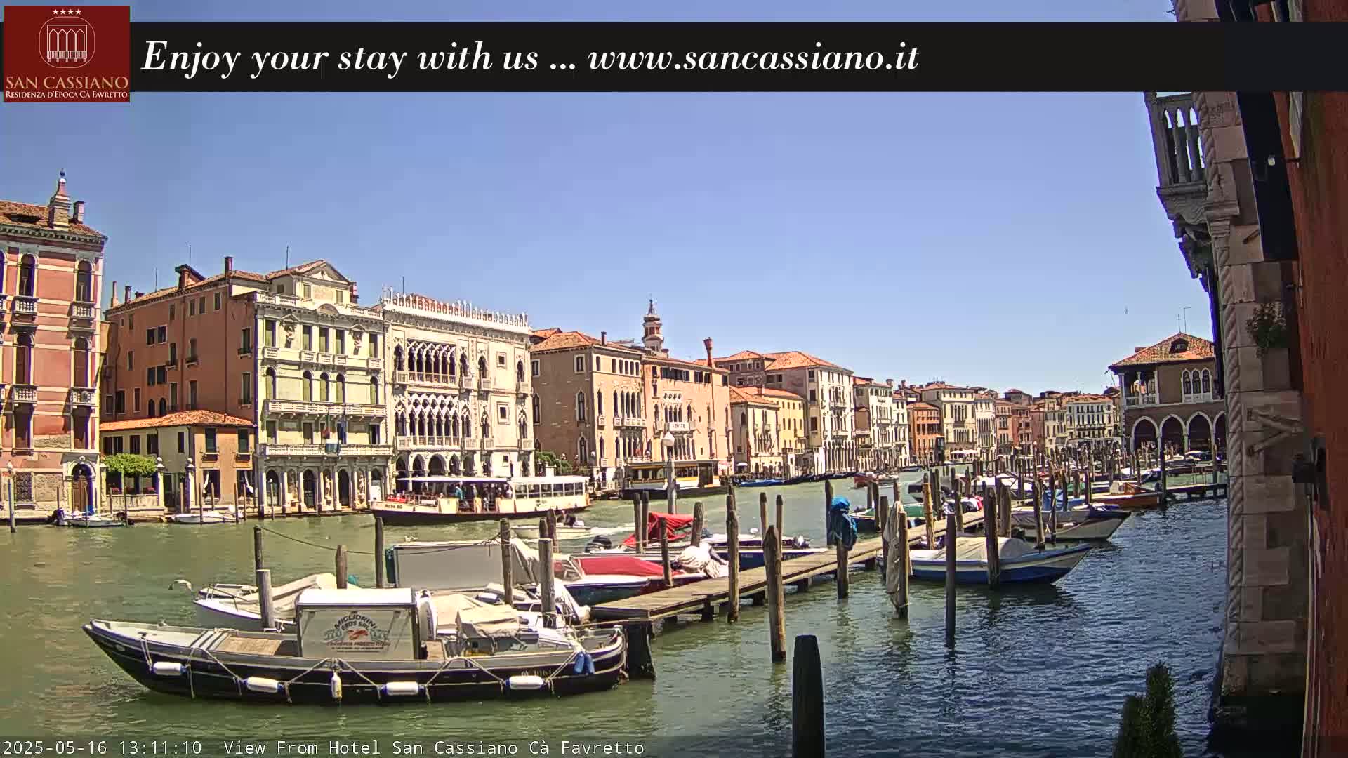 Under a clear blue sky, numerous boats are moored at a dock along a canal lined with historic Venetian buildings.
