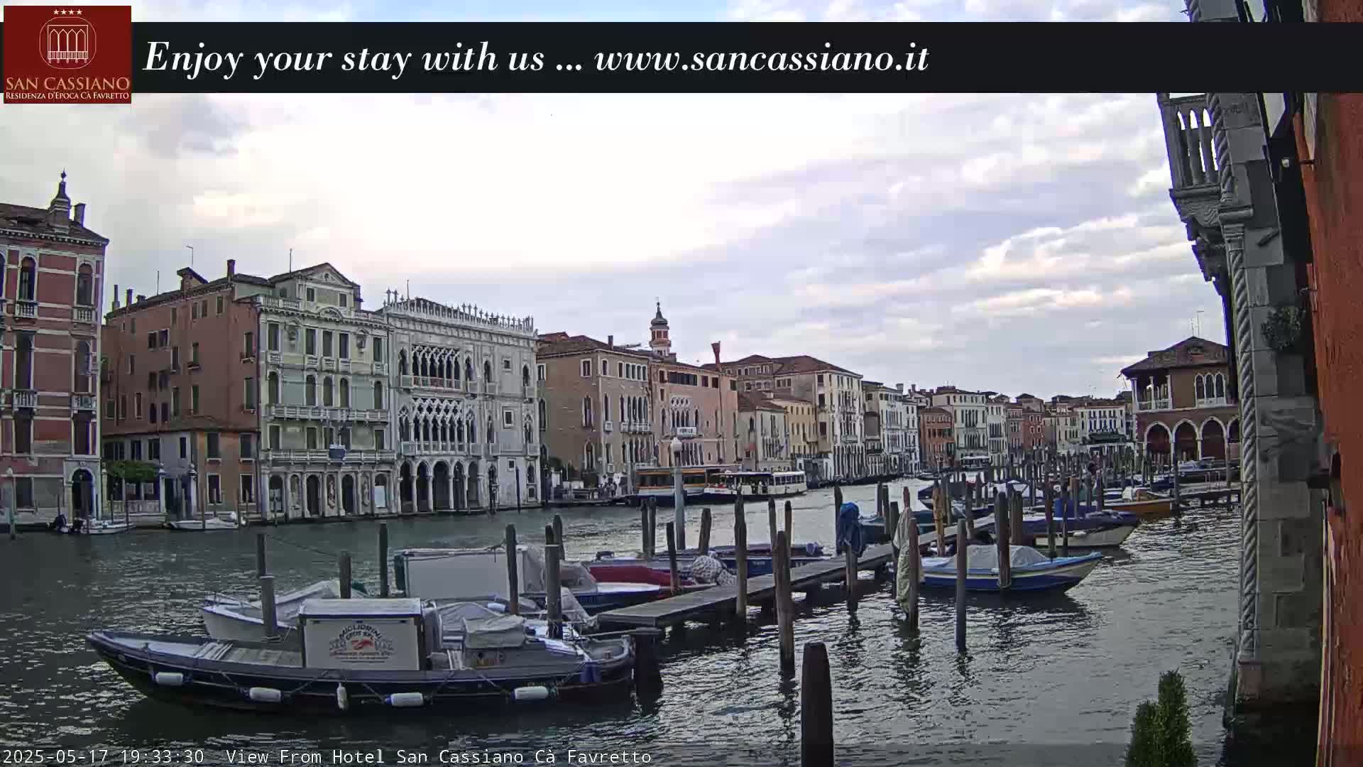 A canal in Venice, under a cloudy sky, is lined with colorful buildings and numerous boats moored at wooden docks.