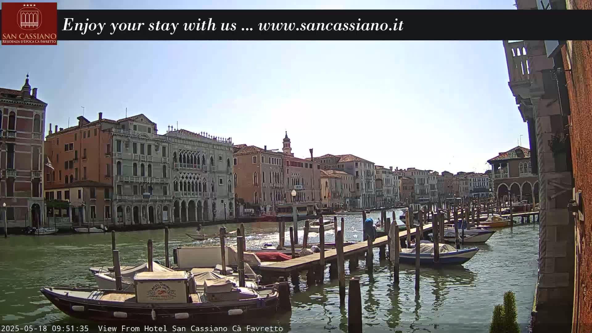 A sunny day in Venice shows a canal lined with historic buildings and several moored boats at a dock.