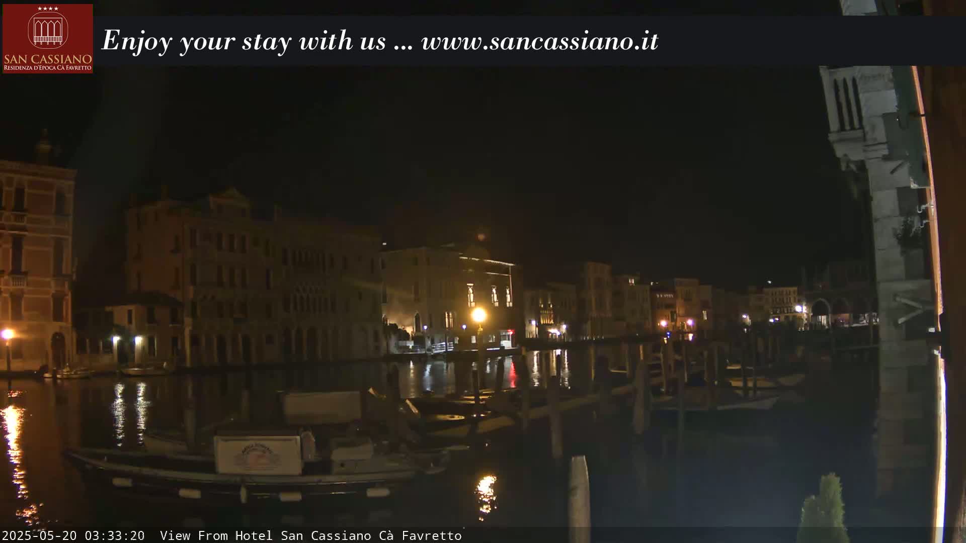 A nighttime view of a canal in Venice, showing illuminated buildings reflected in the dark water, with boats moored alongside a dock.