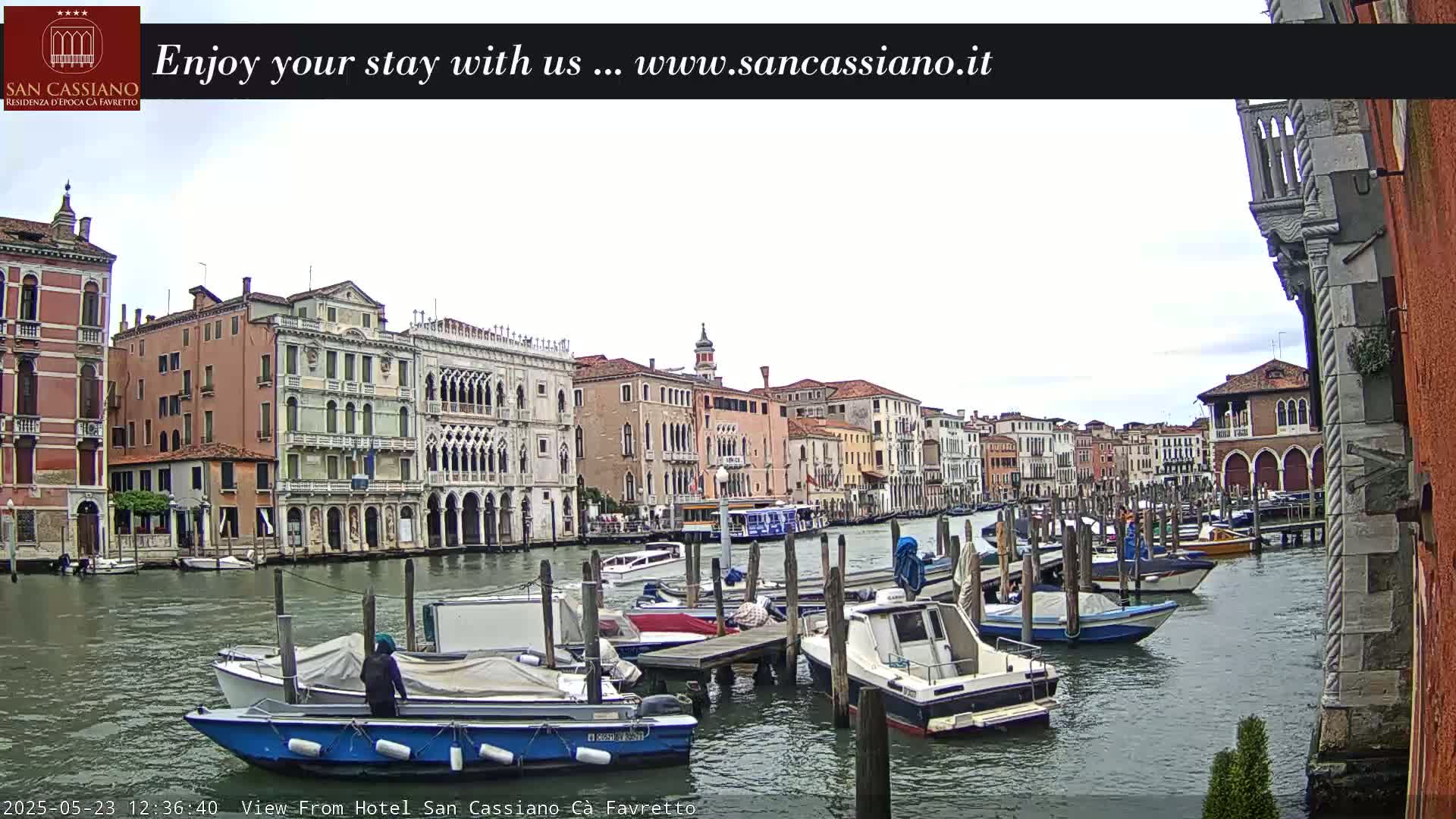 A canal in Venice, Italy, is shown on an overcast day, with numerous boats moored along the waterway and several multi-story buildings lining the banks.