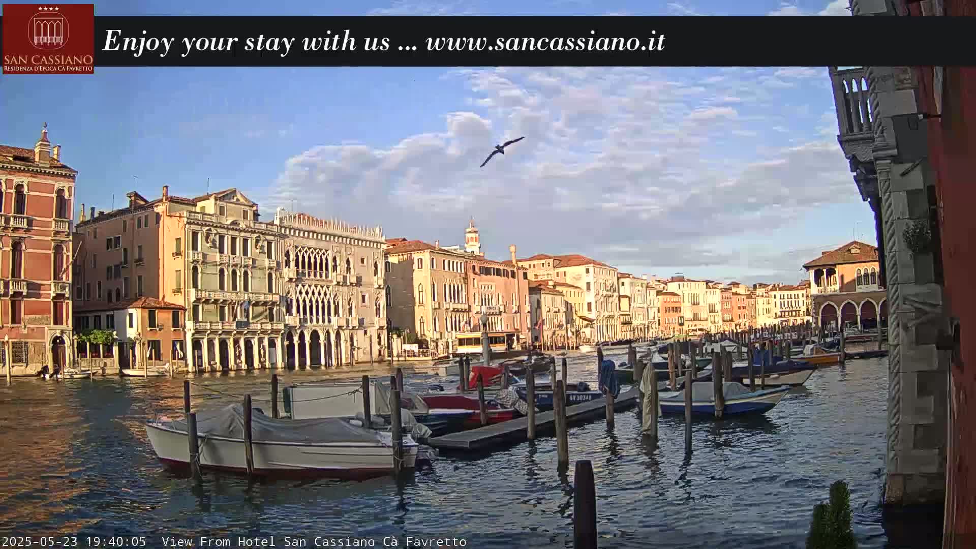 A canal in Venice, Italy, with numerous boats moored along its banks, under a partly cloudy sky.