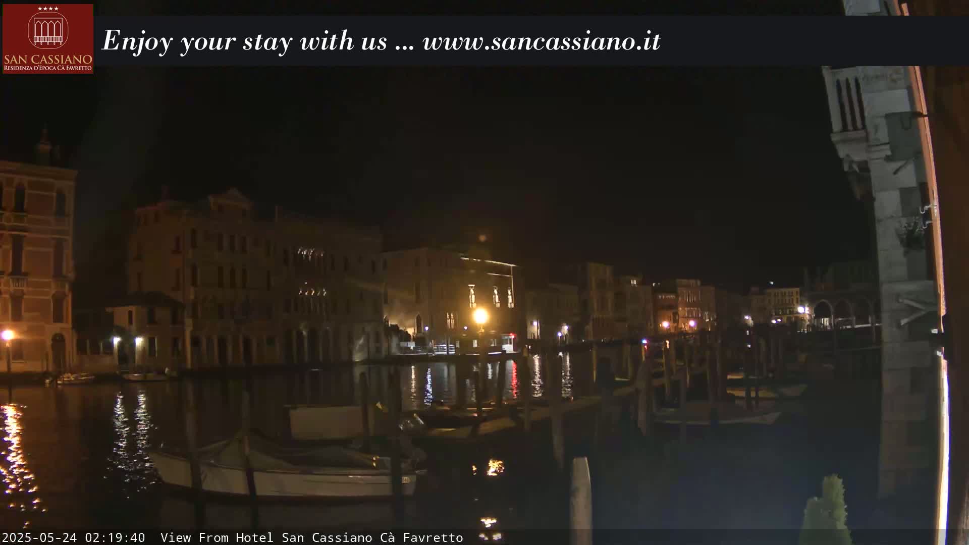 A nighttime view of a canal in Venice, with buildings illuminated along the waterway and several boats moored at the edge.