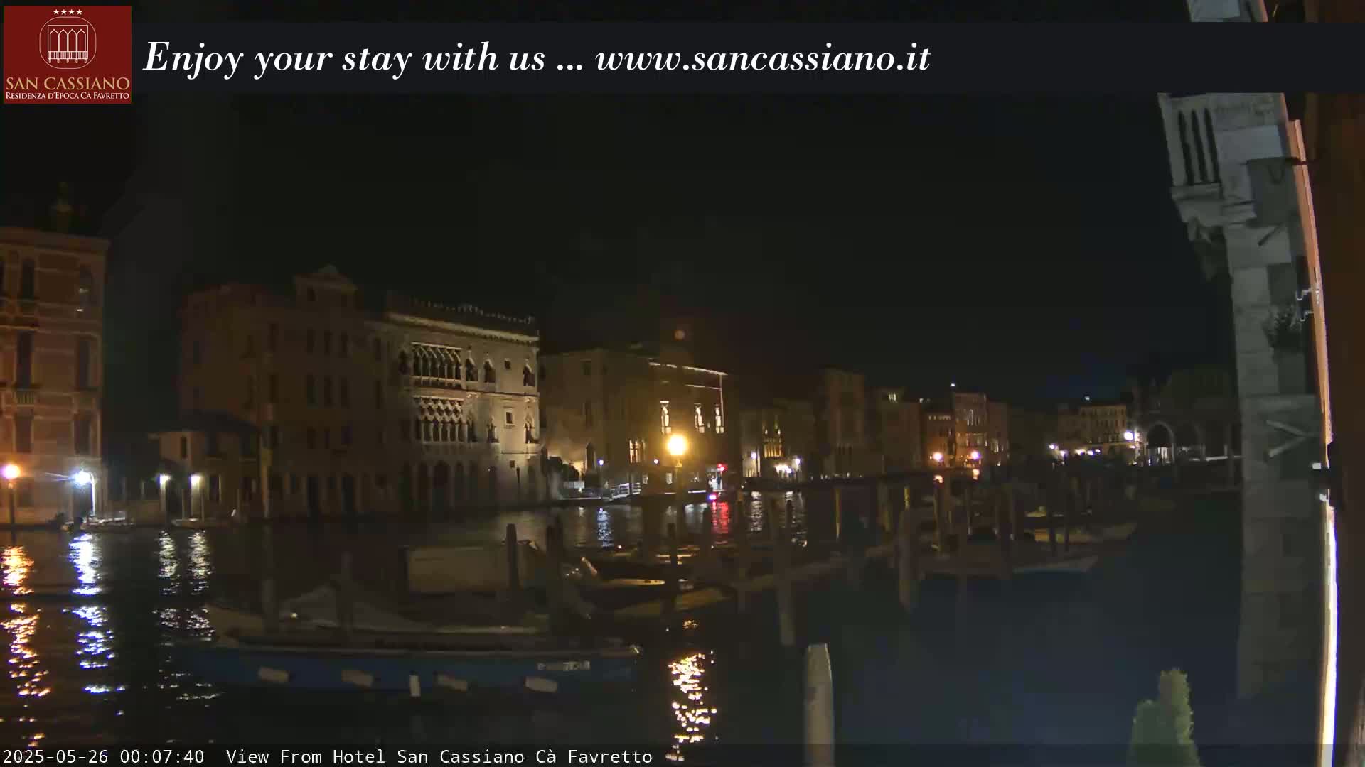 A nighttime view of a canal in Venice, with several buildings illuminated along the waterway and boats moored at the edge.