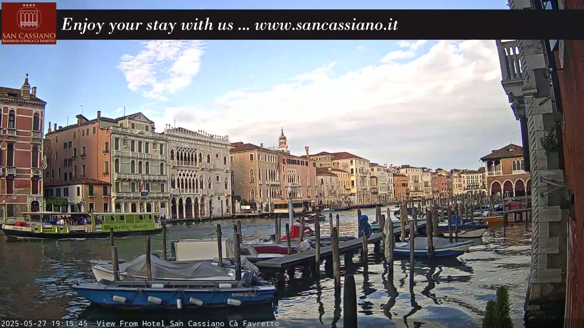 A waterway scene in Venice, under a partly cloudy sky, shows numerous boats moored at a dock in front of several historic buildings.