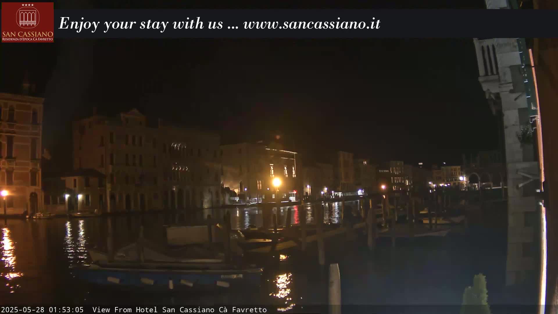 A nighttime view of a canal in Venice, showing illuminated buildings along the waterway and several boats moored.