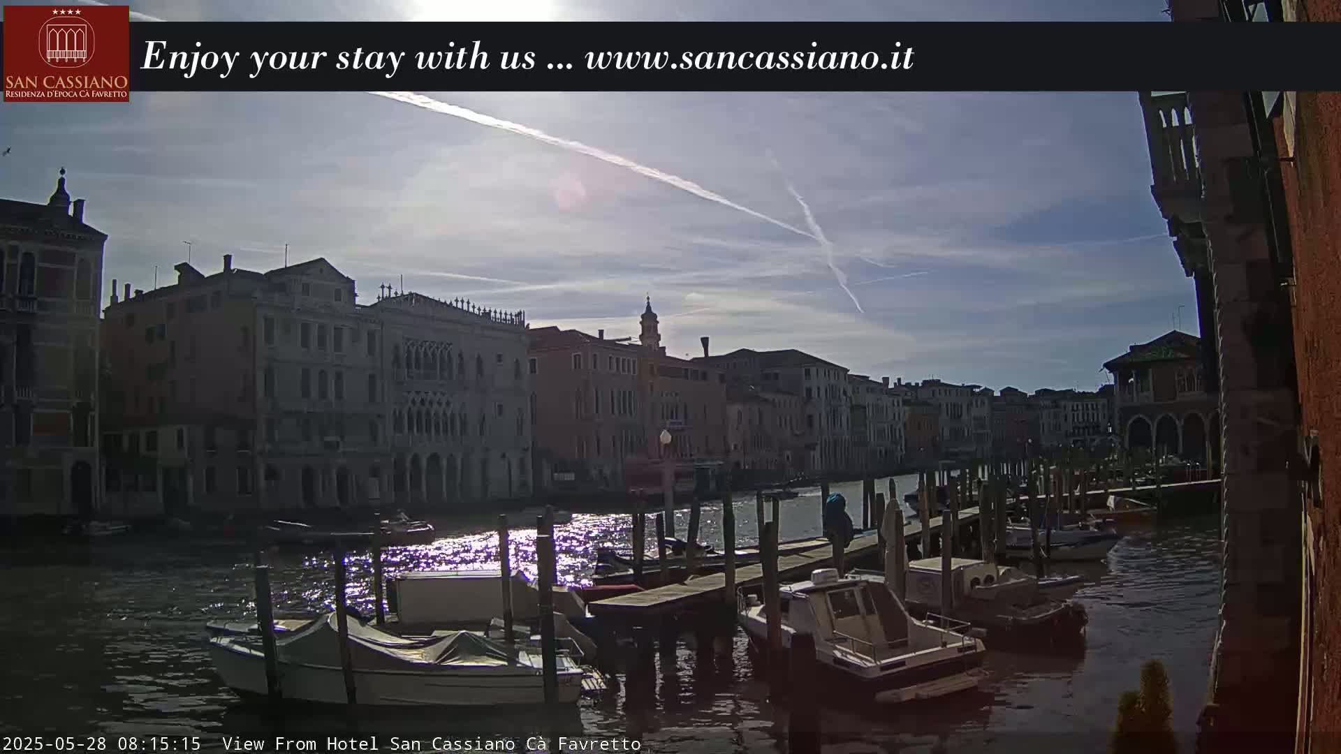 A sunny day in Venice shows several boats moored at a dock along a canal, with buildings lining the waterway under a partly cloudy sky.
