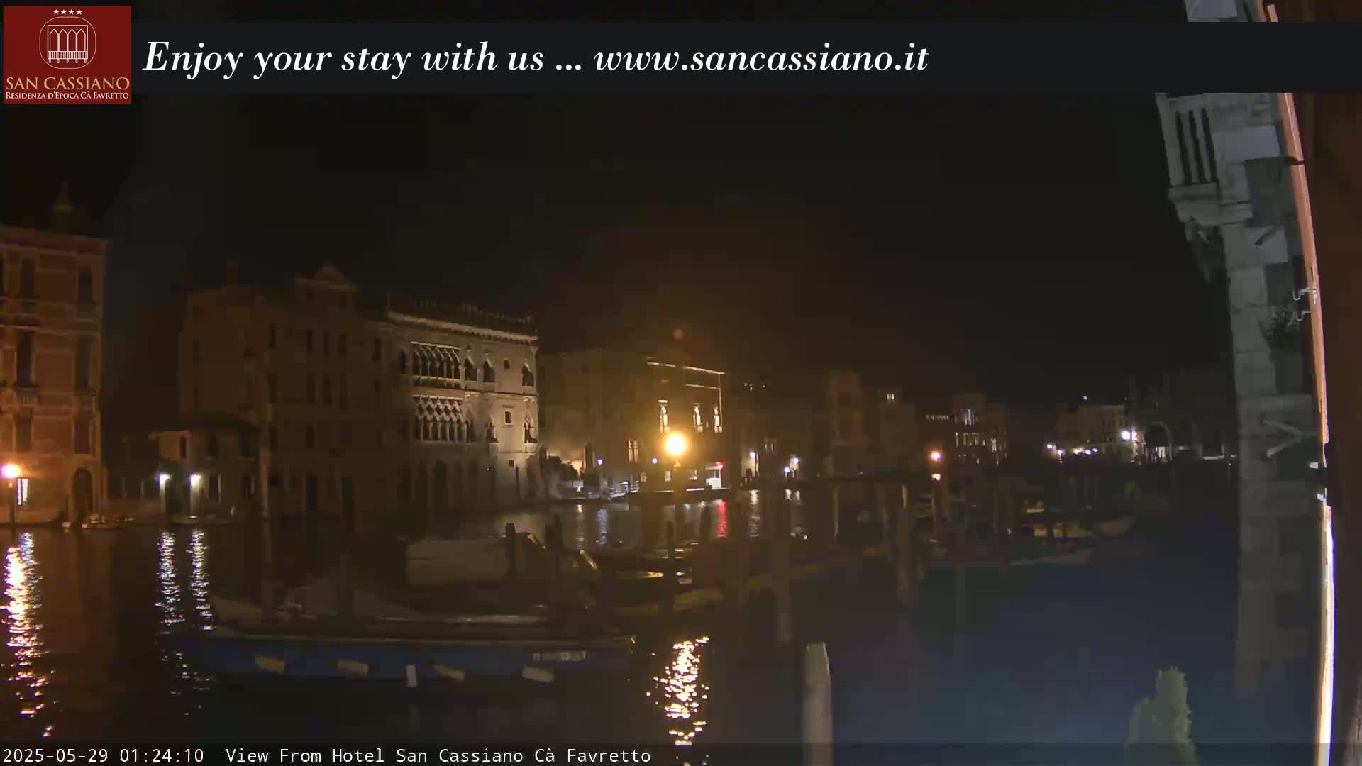 A nighttime view of a canal in Venice, showing illuminated buildings along the waterway and several boats moored.