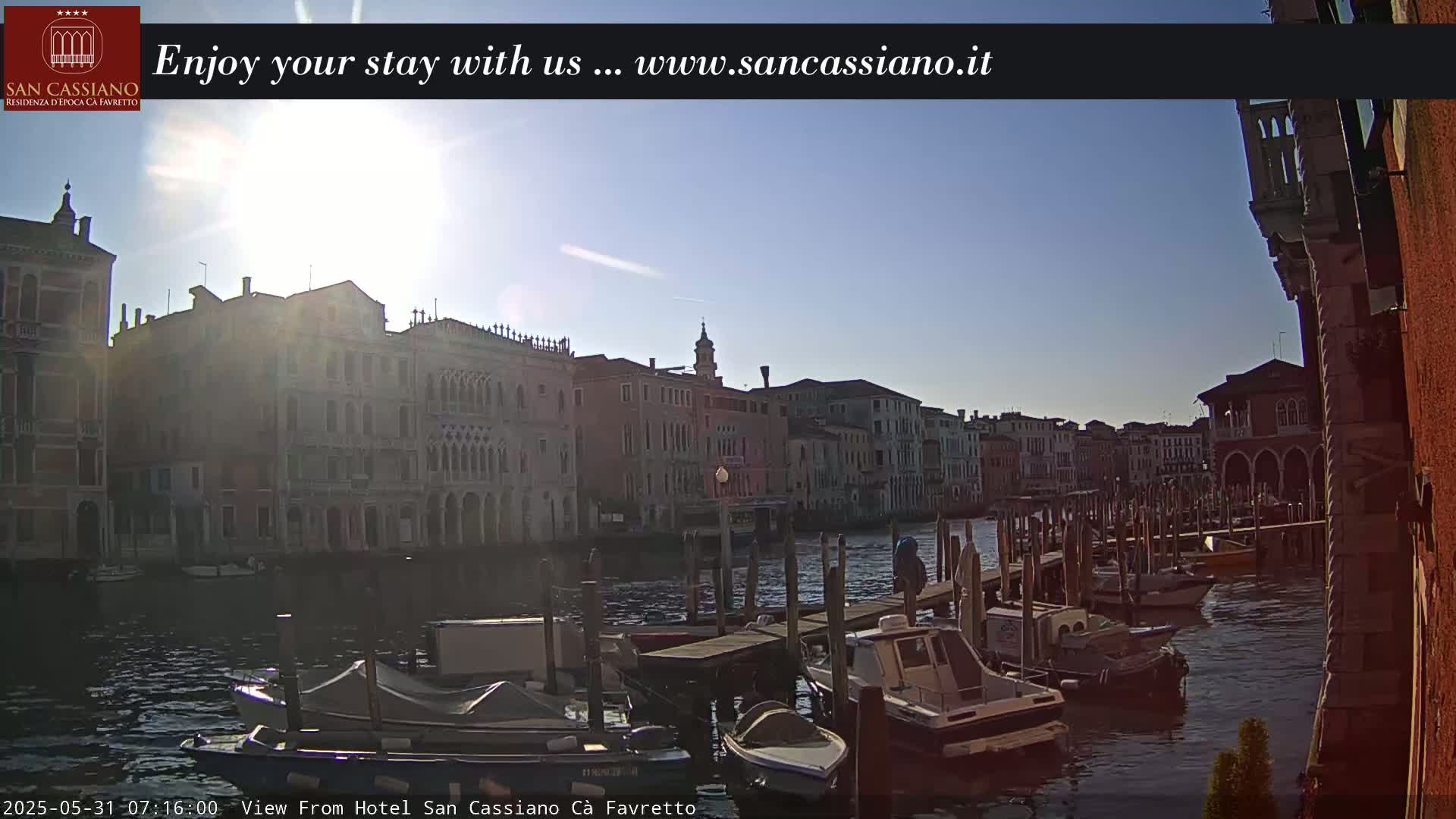A sunny Venetian canal scene shows several docked boats and buildings lining the waterway.